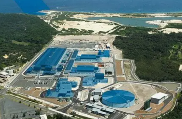 Aerial view of an industrial facility near a body of water, with multiple large blue buildings and surrounding roads and greenery.