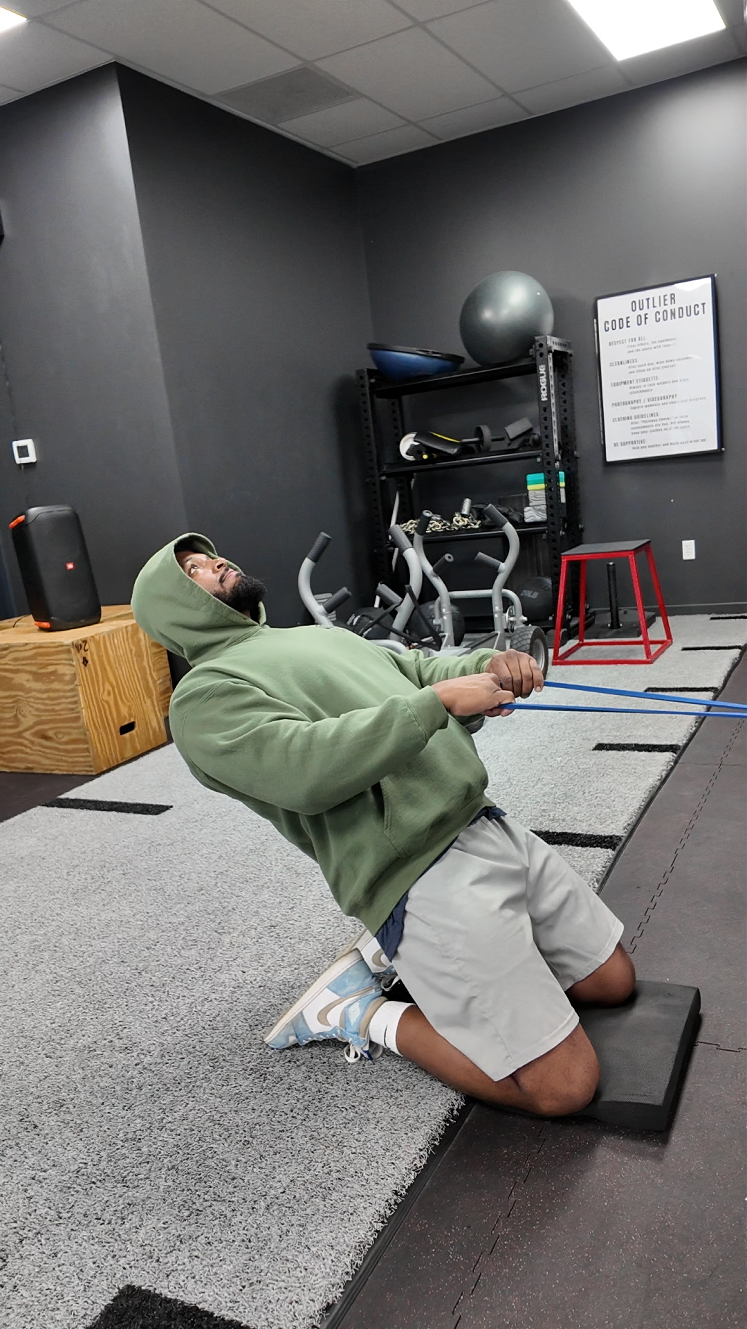 A man in a green hoodie kneeling on a black mat doing resistance band exercises in a gym. The gym has dark gray walls, exercise equipment, and a framed 'Outlier Code of Conduct' on the wall.