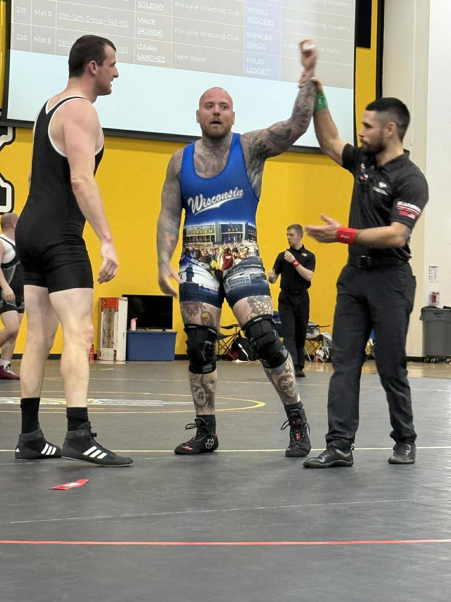 A male wrestler wearing a blue 'Wisconsin' singlet is being declared the winner by a referee, who is raising his hand. Another wrestler in black and a person in black clothing are standing nearby. The scene takes place in a wrestling gymnasium with a large screen displaying brackets or results in the background.