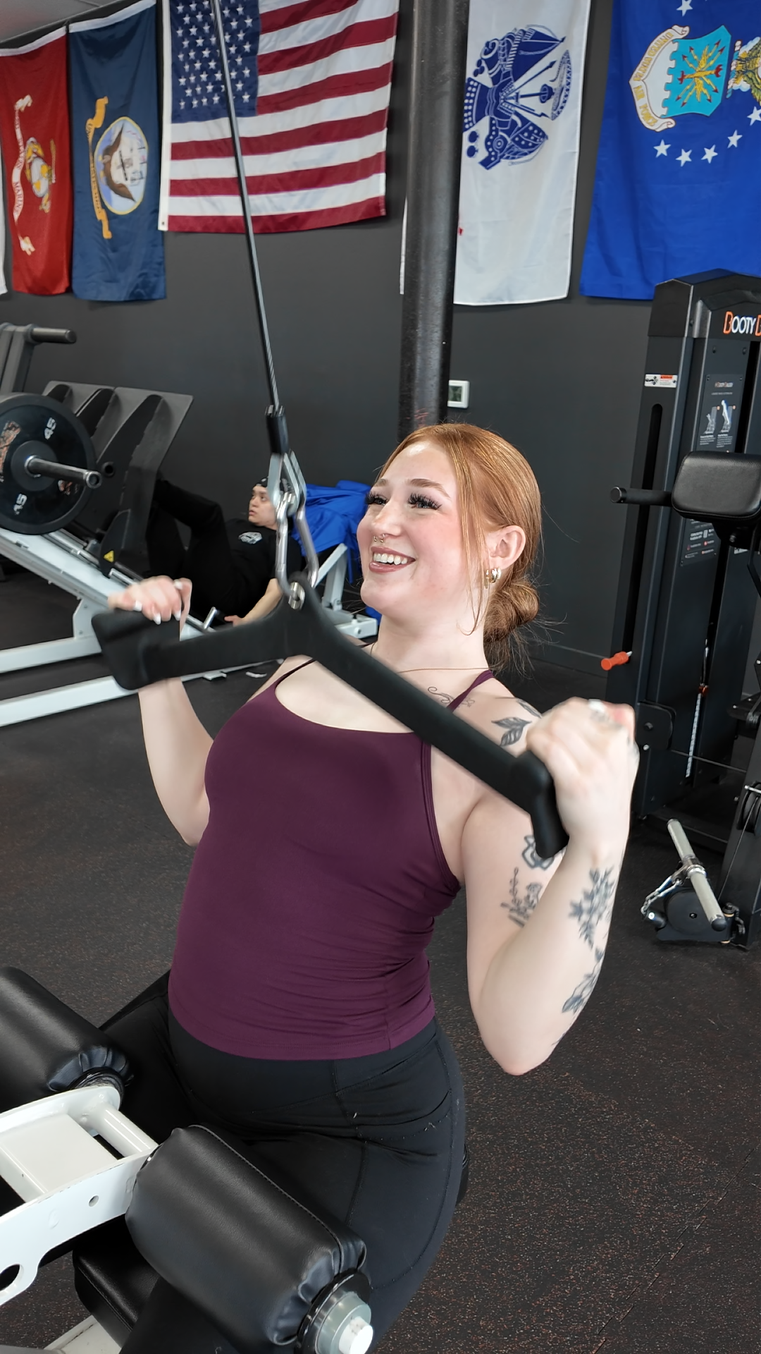 Young woman with tattoos exercising on a shoulder press machine at gym, smiling, with American and military flags hanging on the wall in the background.