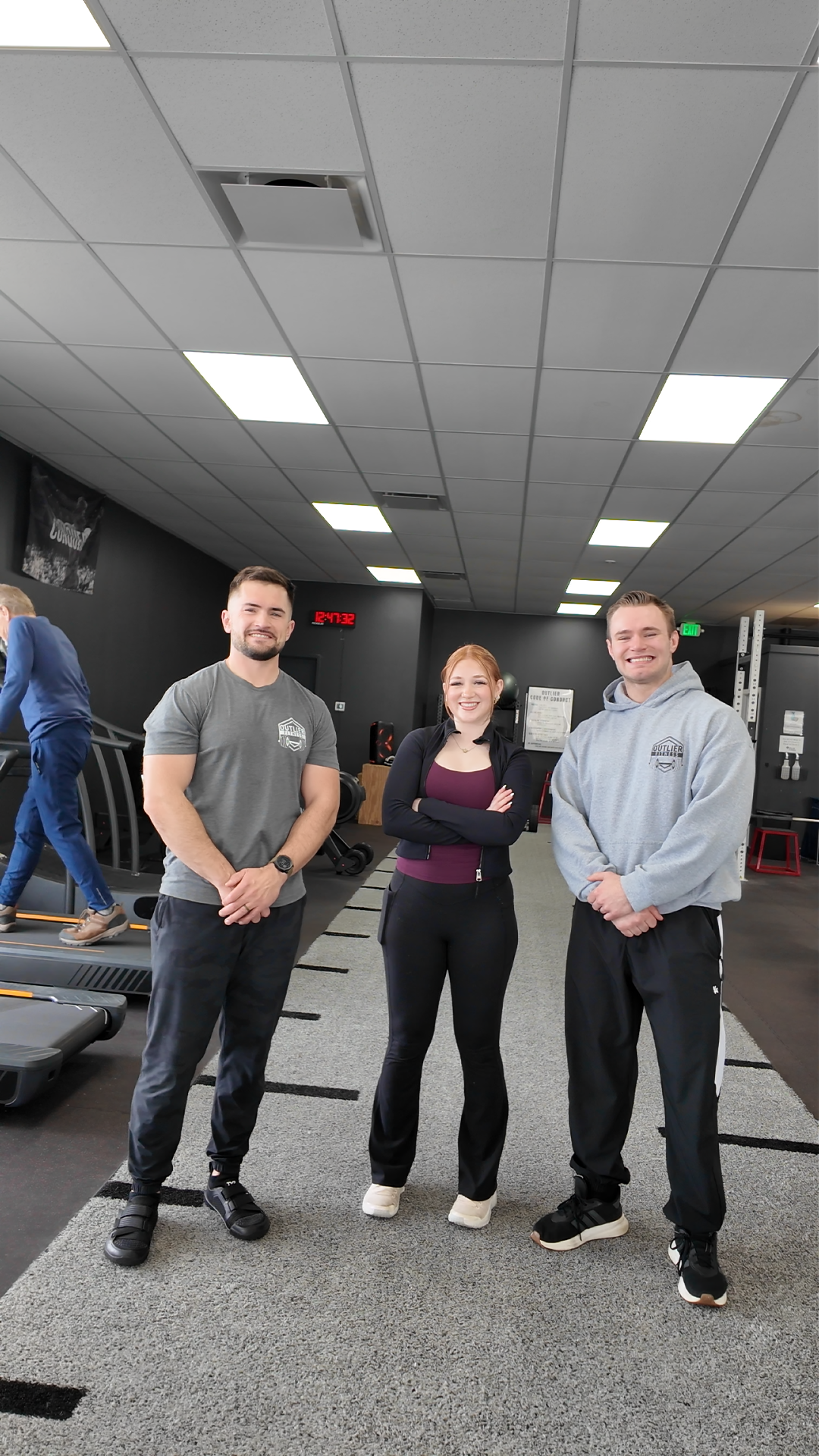 Three smiling young adults standing together in a gym, with exercise equipment and a treadmill in the background.