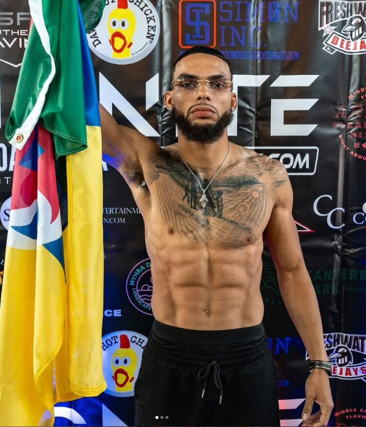 A shirtless man with glasses and a beard standing in front of a black background with various logos, holding a flag and displaying a serious expression.
