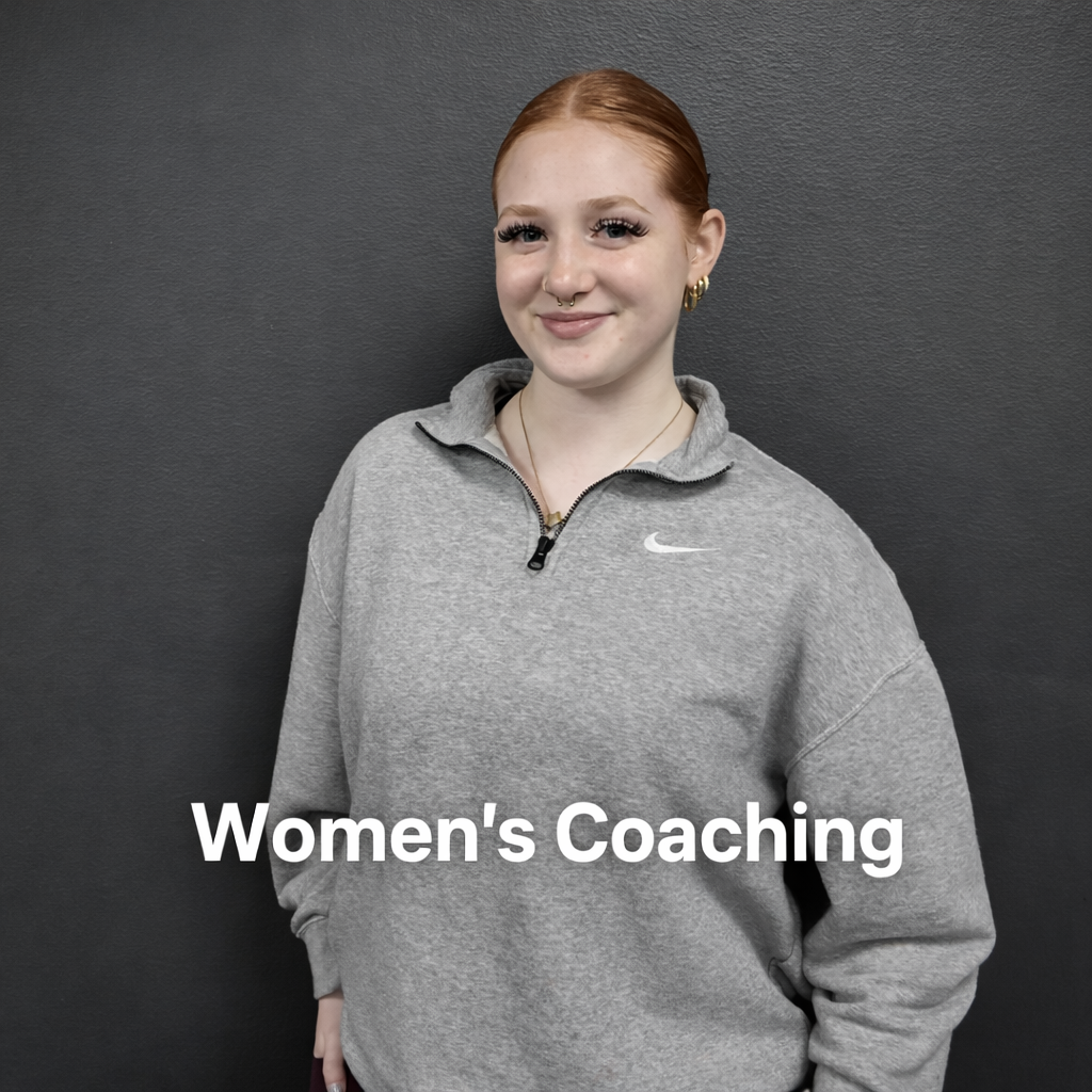 A woman with red hair, wearing a gray Nike zip-up sweatshirt, standing against a gray wall with a smile.