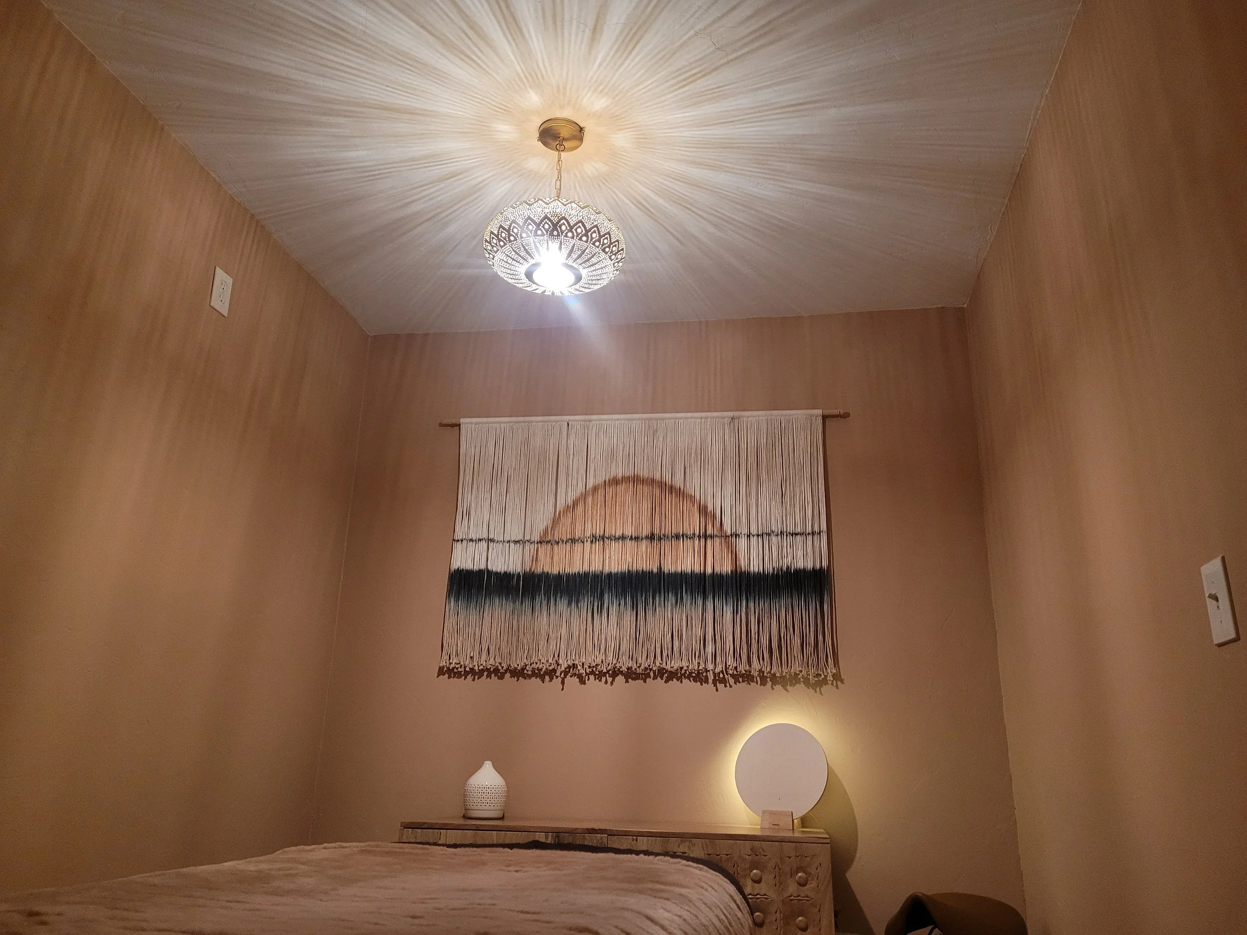 A bedroom with beige walls and a textured ceiling, lit by a modern ceiling light. A woven wall hanging featuring a circle and horizontal lines hangs above a wooden dresser. On the dresser are a white decorative vase and a round lamp with a yellow glow.