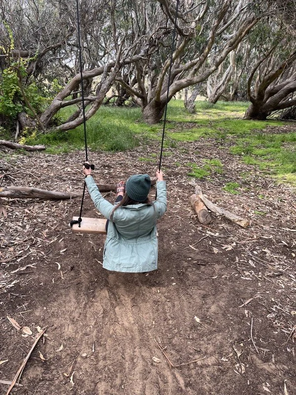 A child sitting on a wooden swing in a wooded area with leafless trees and green grass.