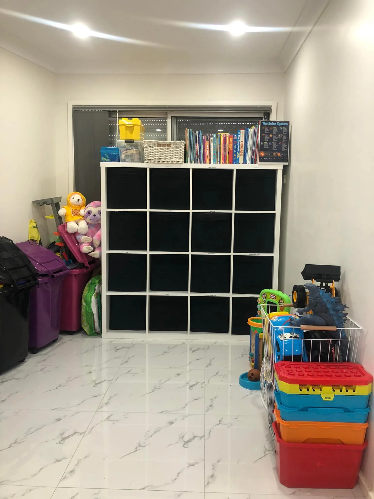 Room with children's toys and storage bins, black cube shelving unit, books, and toys on the right, with a window behind the shelf and a white marble tile floor.