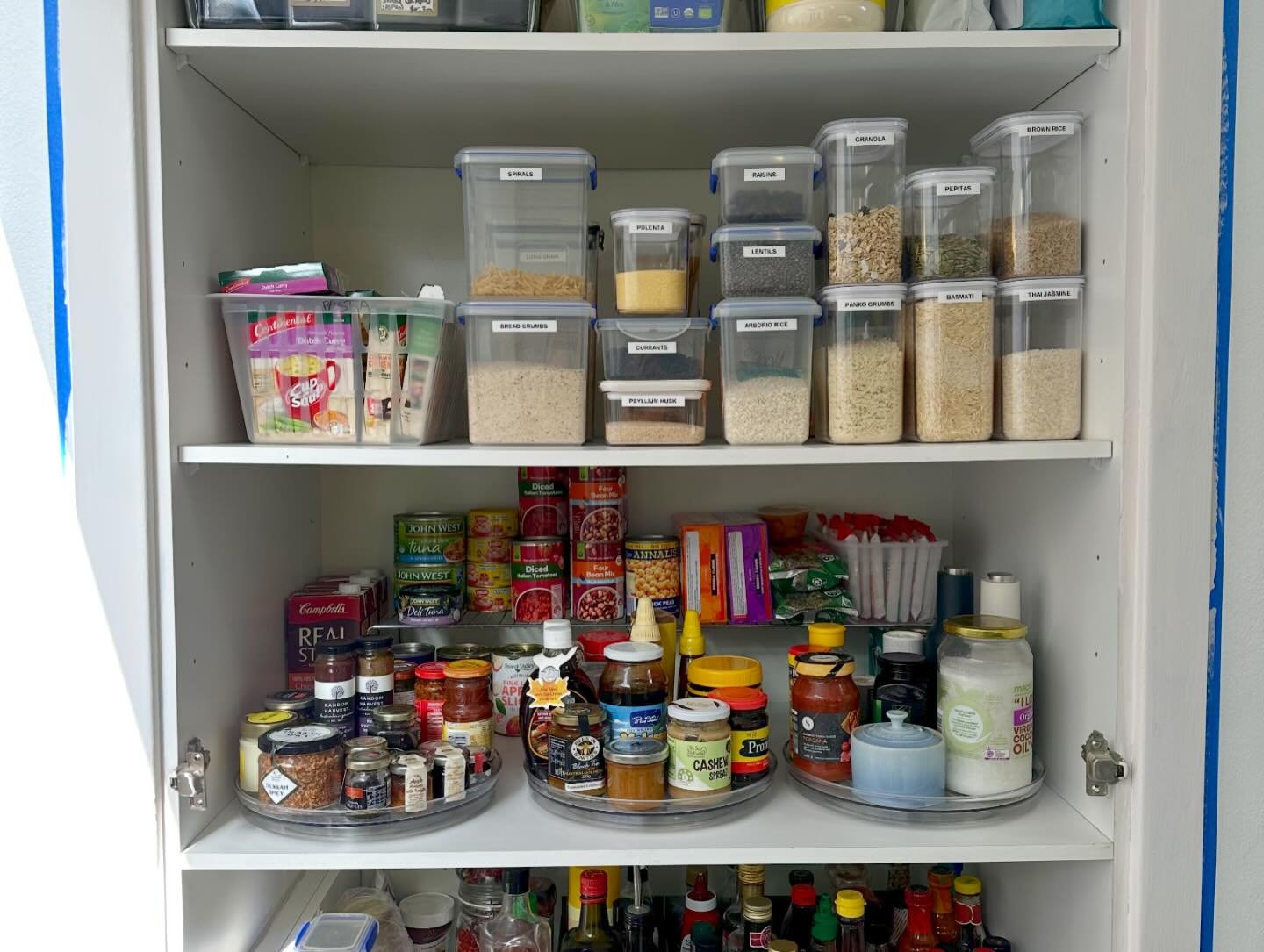 Kitchen pantry cabinet filled with various canned goods, spices, jars, and food containers, organized on shelves.