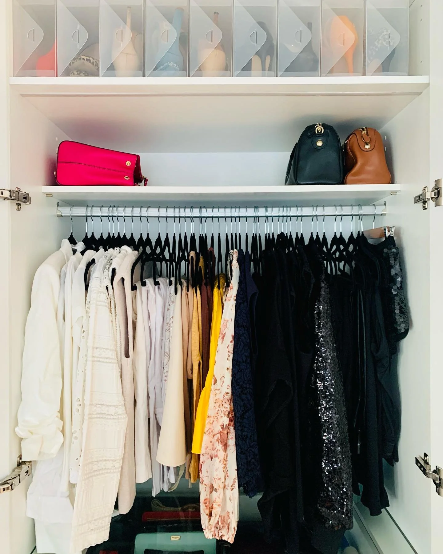 Inside of a closet with hanging clothes and three handbags on a shelf, with shoe boxes on a top shelf.