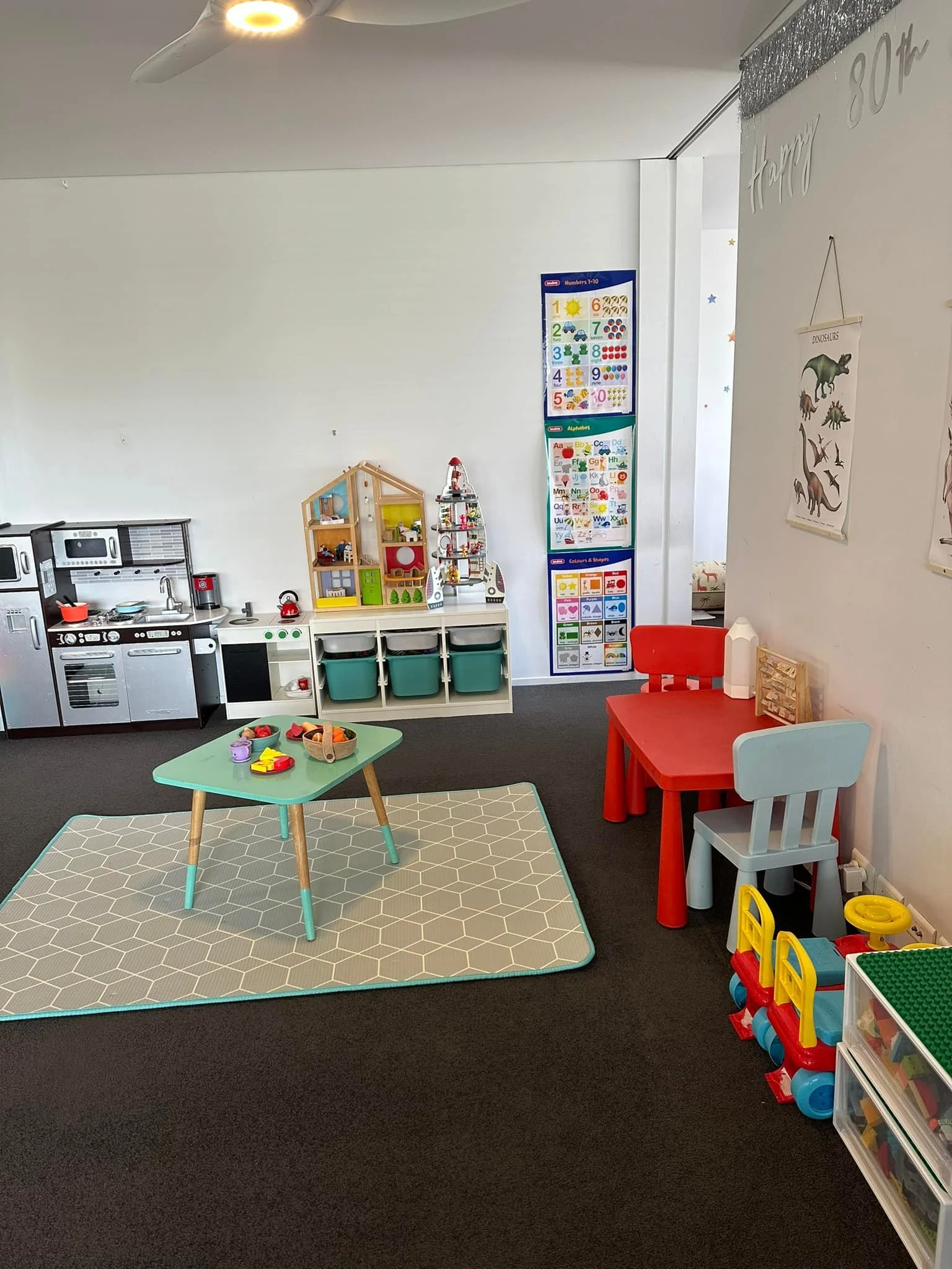 A children's playroom with toy kitchen, small table with toys, a red and a gray children's chair, and educational posters on the wall. The room has a dark carpeted floor and a white wall.