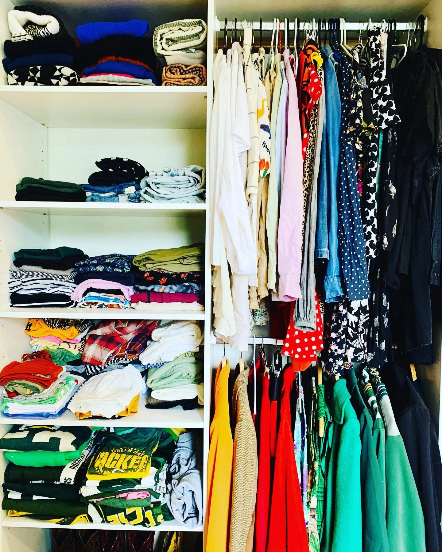 A closet with white shelves on the left filled with folded clothes in various colors, and a hanging rod on the right with shirts, dresses, and jackets in different patterns and colors.