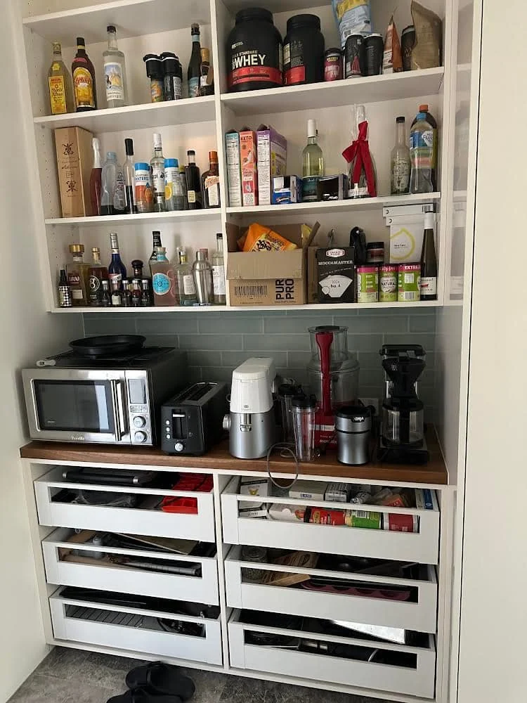 Kitchen cabinet with open shelves filled with bottles, jars, and food items on top and small kitchen appliances like microwave, toaster, and blender on the countertop below.