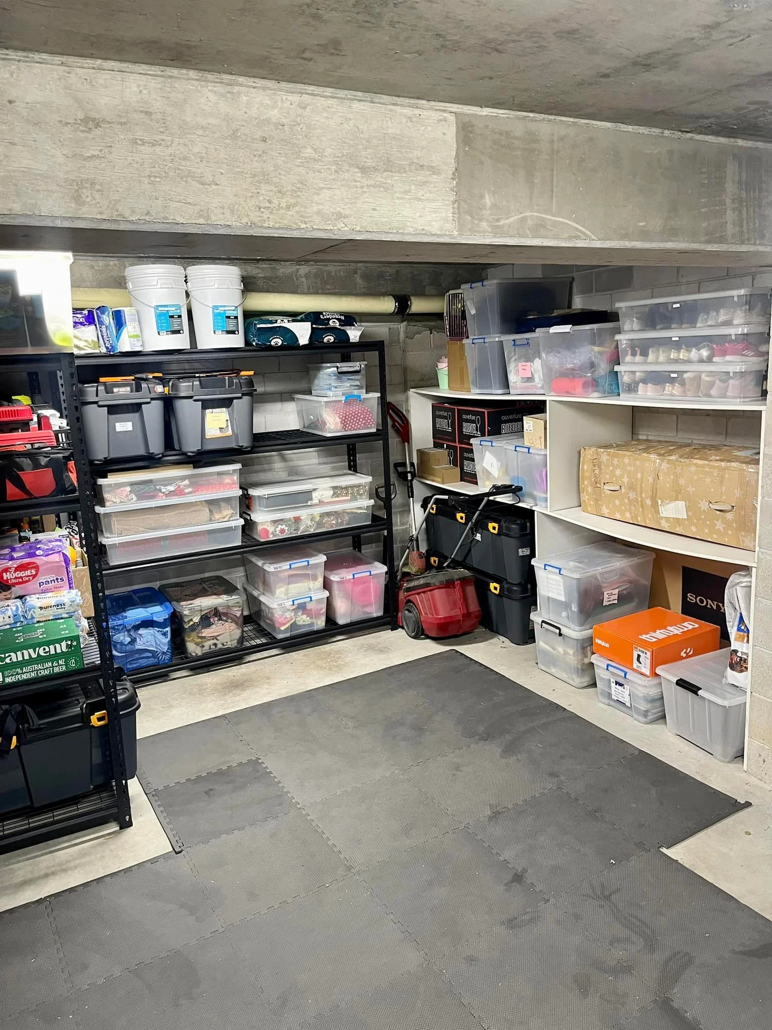 A garage storage room with shelves filled with plastic bins, boxes, and tools, and a small workspace on the floor.