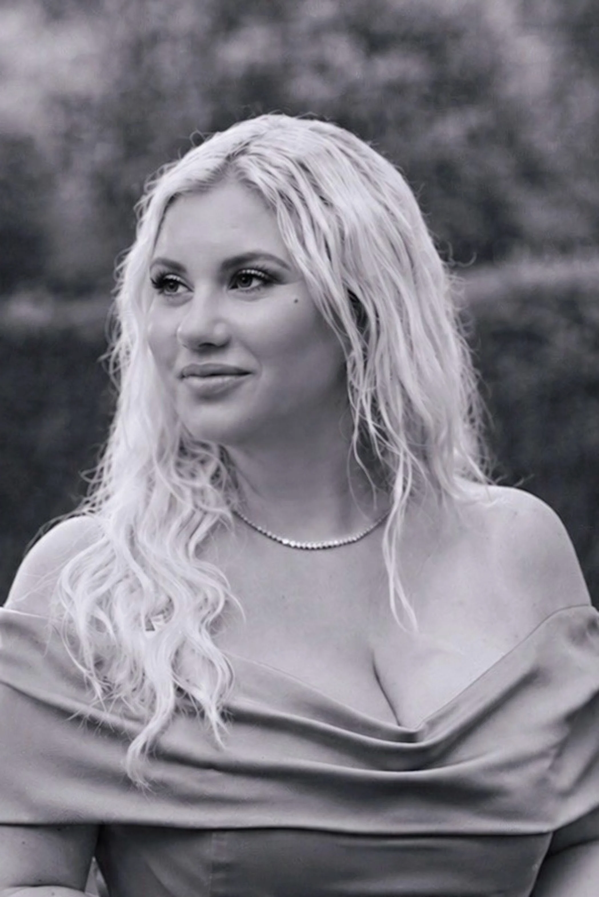 Black and white photo of a woman with long, wavy, light-colored hair looking slightly upward and to her left, wearing a silver necklace and a loose-fitting gray top, outdoors with blurred background. Author M.S. Donovan portrait.