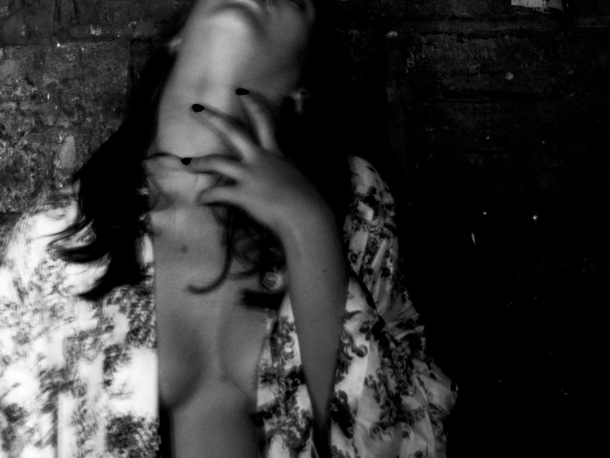 Black and white photo of a person with curly hair, partially turned, wearing a patterned shirt, against a dark textured background.