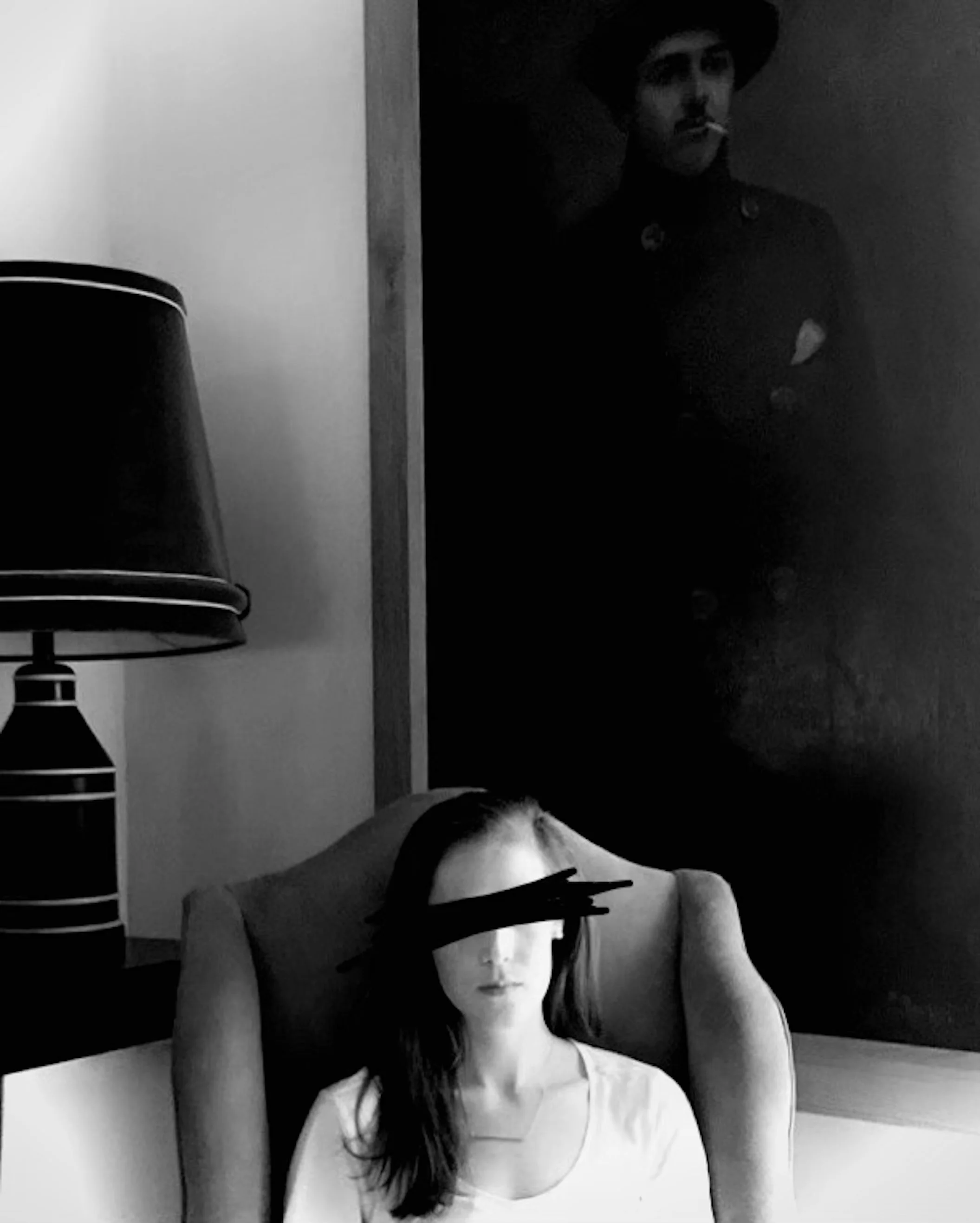 Black and white photo of a woman with long dark hair, sitting in front of a framed portrait of a man with a mustache and hat. A lamp is visible on the left side of the image.