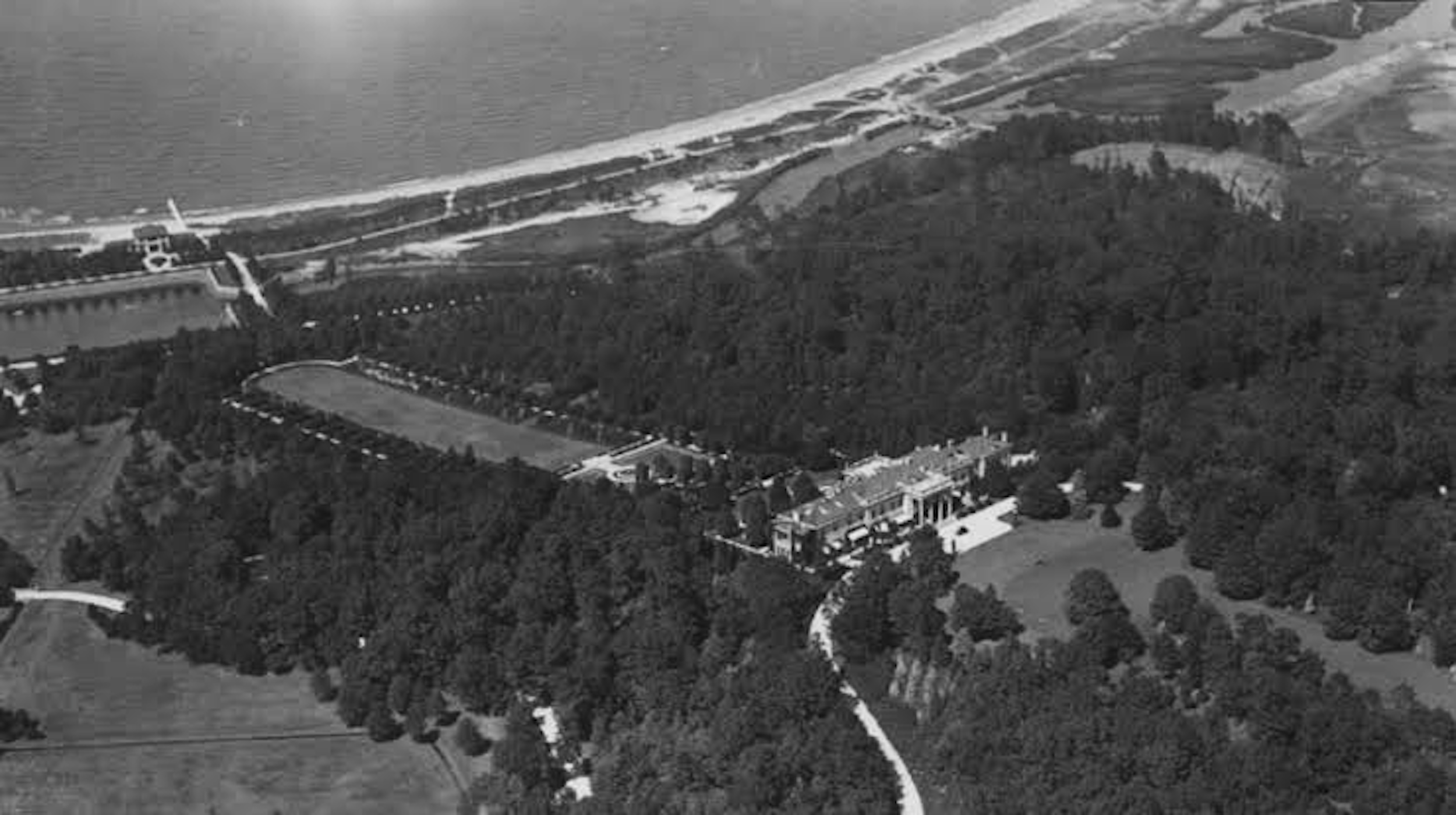 Aerial black and white photo of Locust Valley, NY coastal area with a beach, a body of water, trees, and buildings surrounded by greenery.