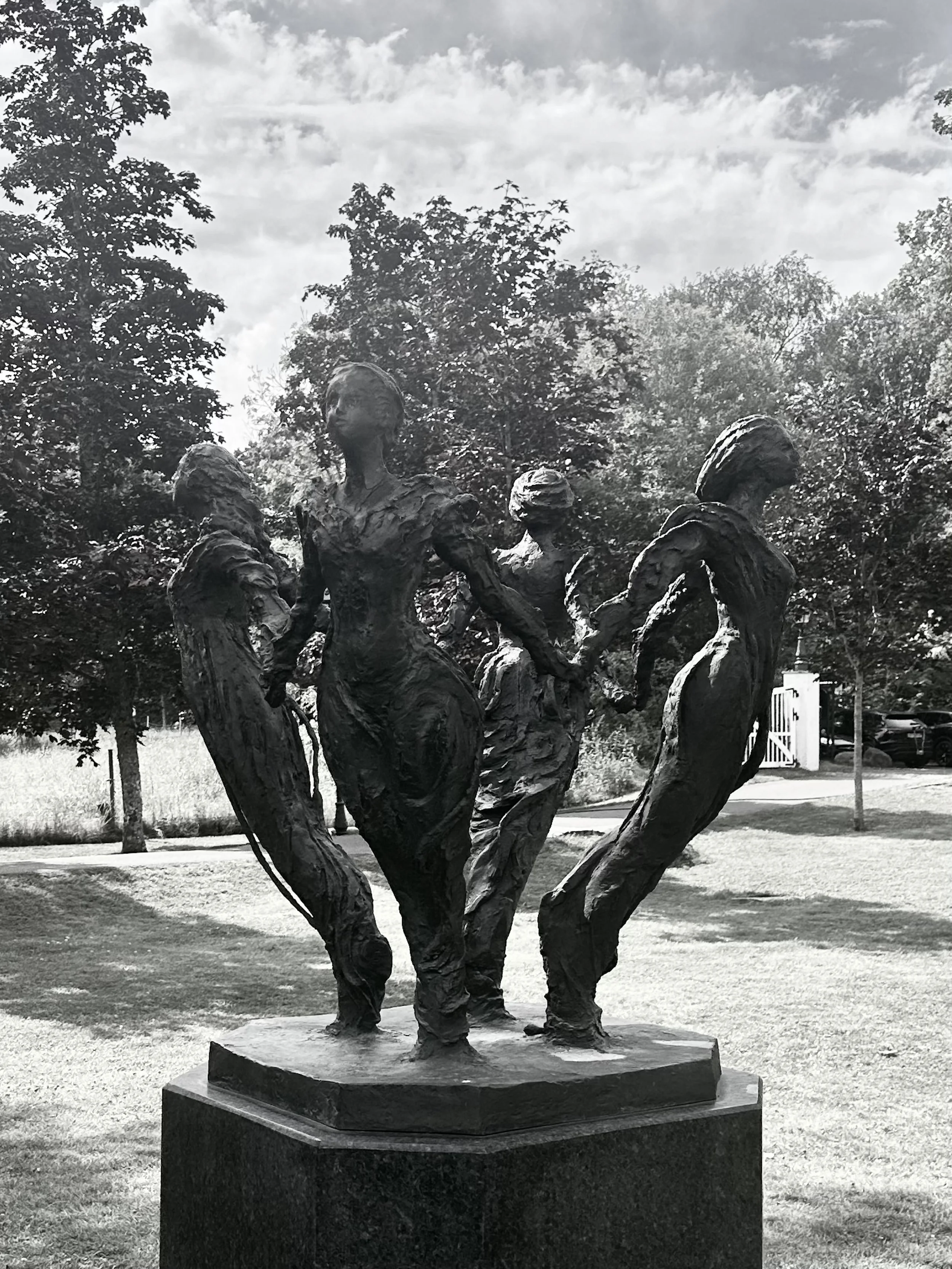 Bronze sculpture of a group of women holding hands, standing together outdoors on a pedestal, with trees and a partly cloudy sky in the background - inspiration for Âmes de la Brume. engø gård - sculpture by Nina Sundoye
