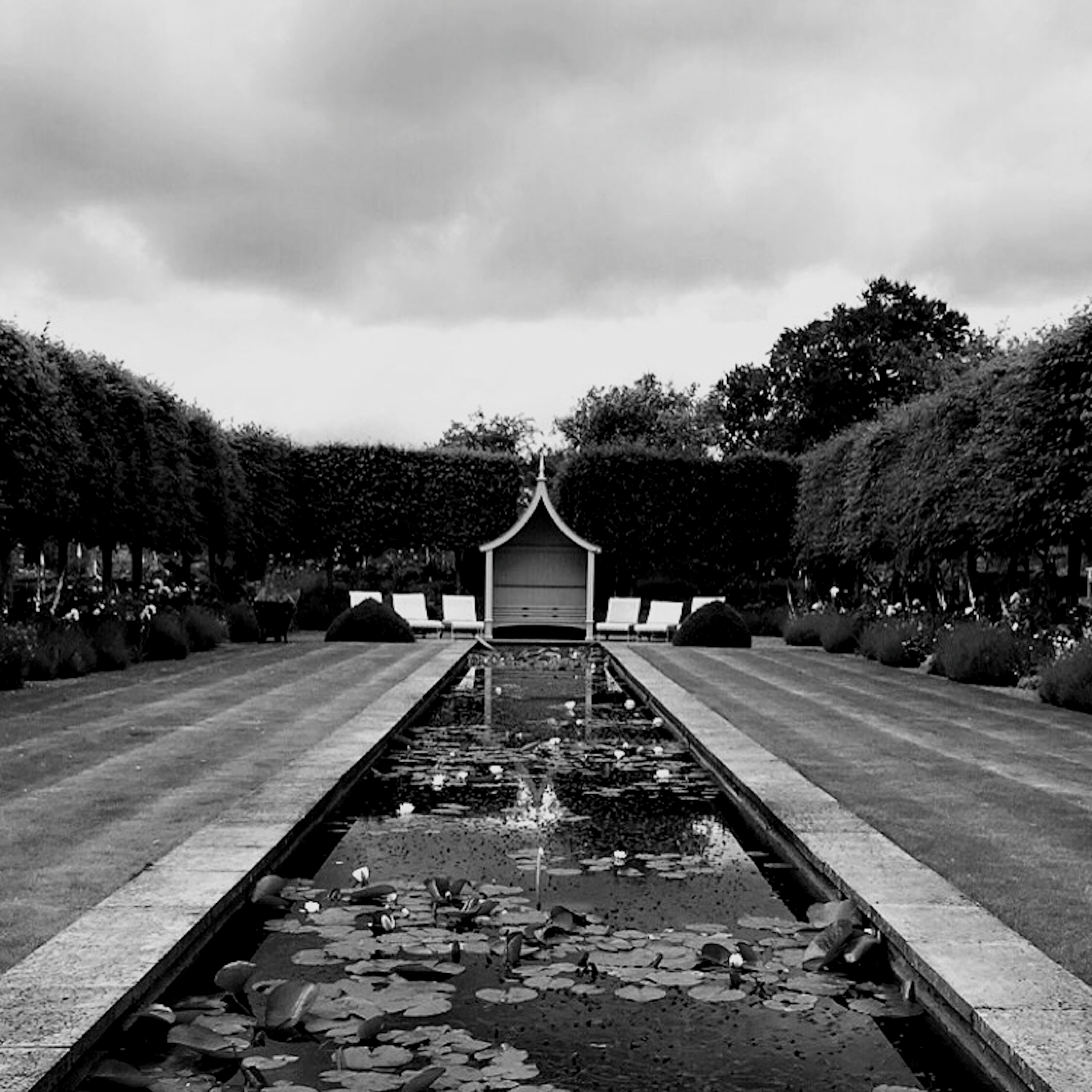 Black and white photo of train tracks leading to a small pavilion or shelter surrounded by trees, with a cloudy sky above.