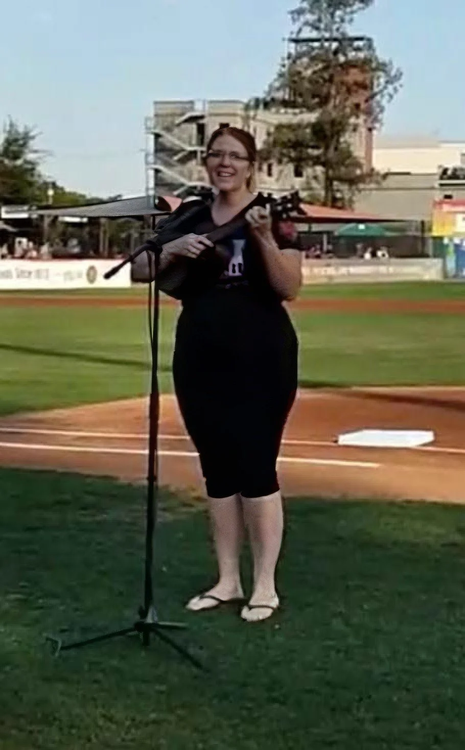 A woman standing on a baseball field holding a ukulele, smiling, wearing glasses, a black shirt, and flip-flops.
