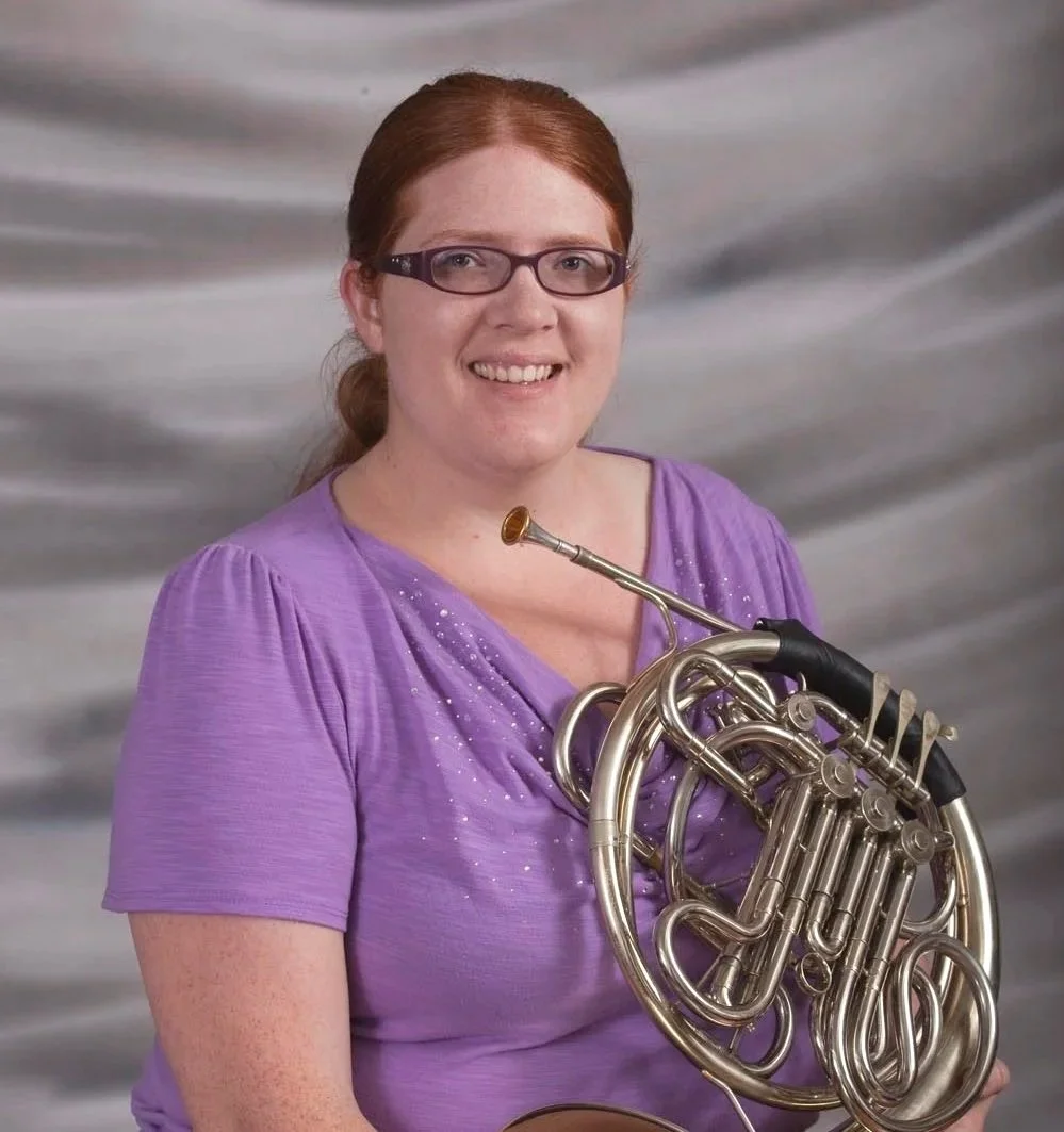 A woman with red hair and glasses holding a French horn, wearing a purple top, standing in front of a gray, marbled background.