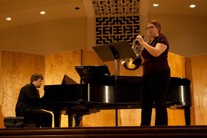 A woman playing French hron and a man playing piano on stage during a performance.