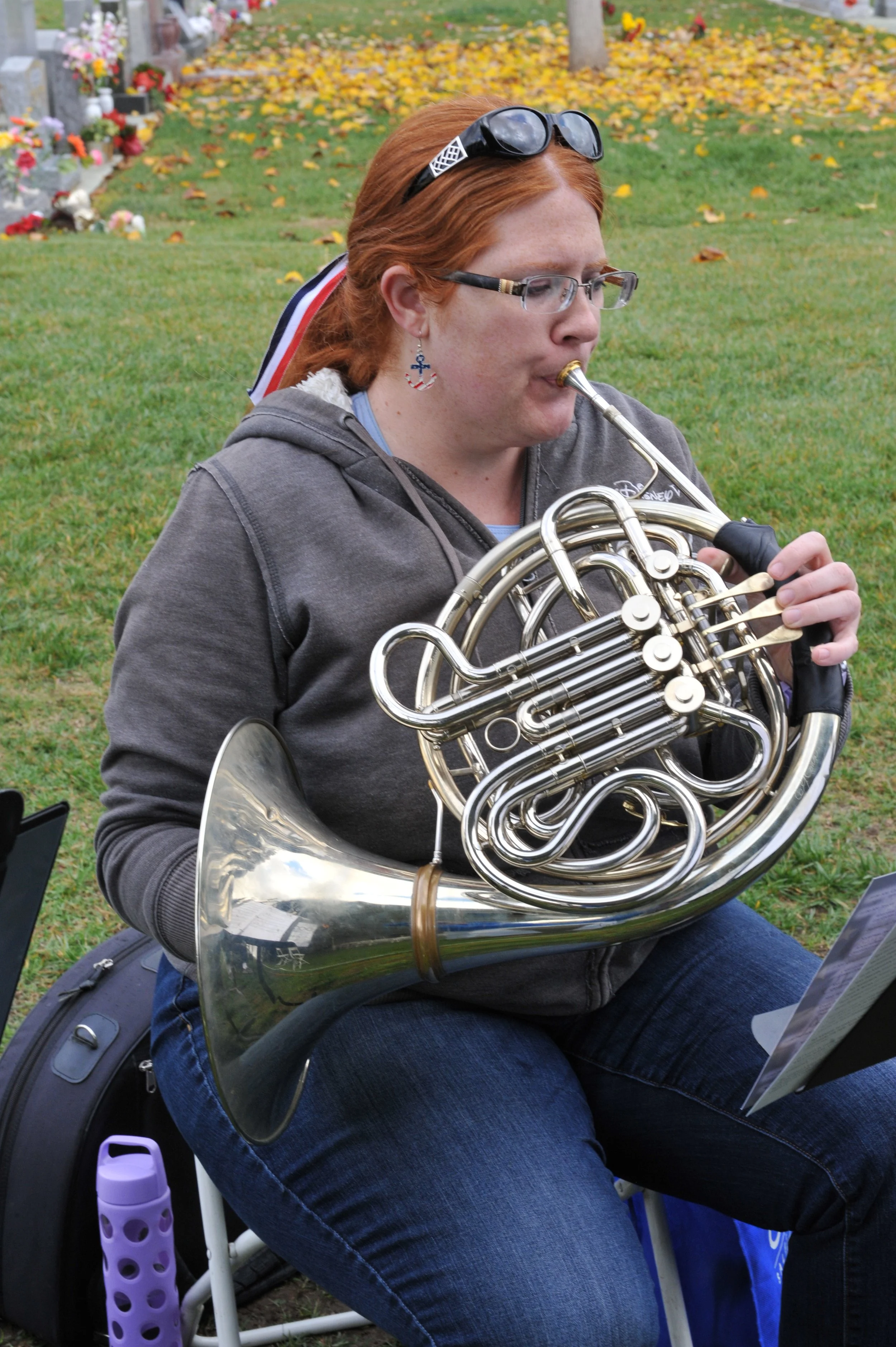 A woman with red hair, glasses, and patriotic earring playing a French horn outdoors in a grassy area near a cemetery with fall leaves.