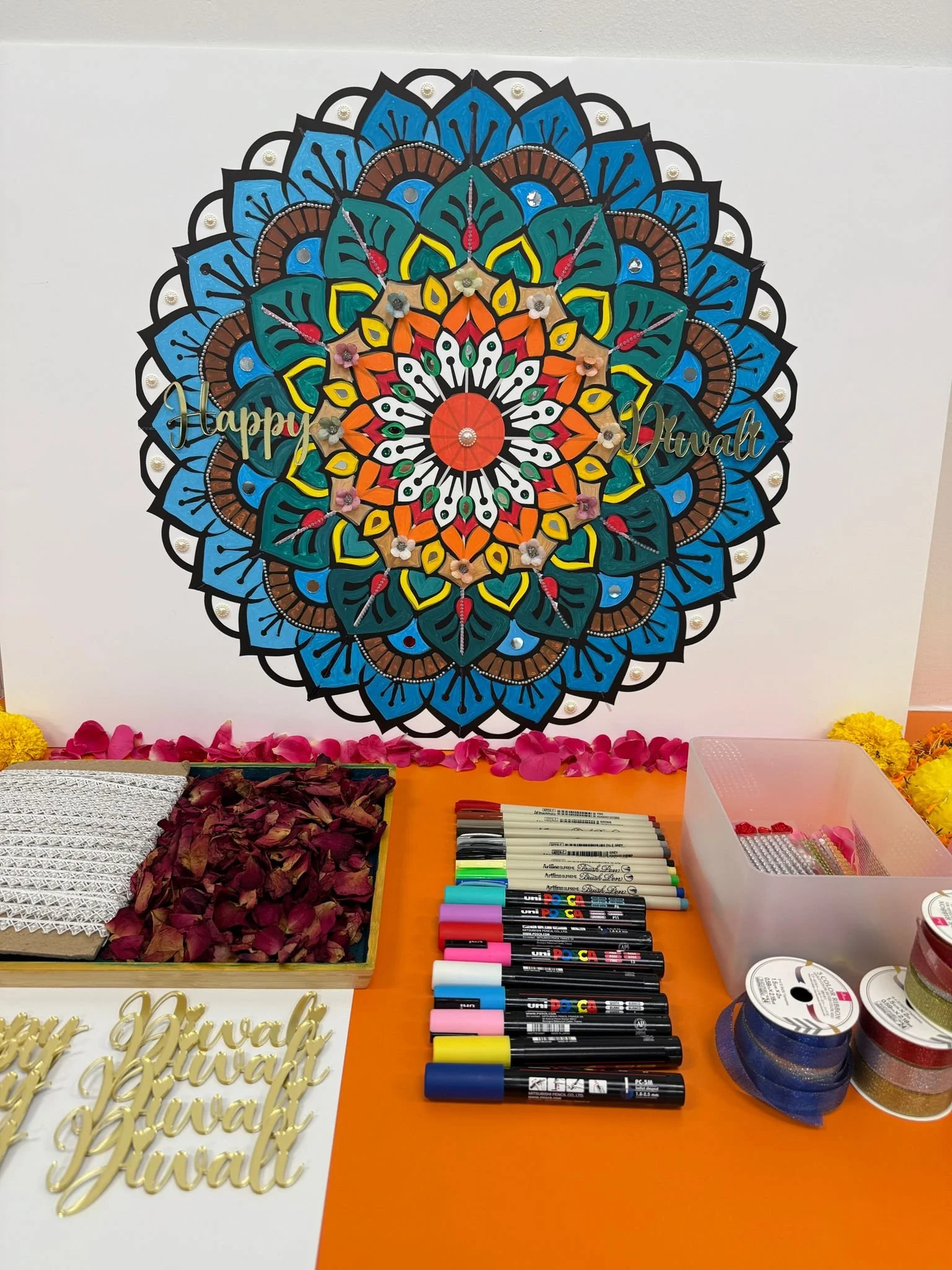 Colorful handmade decoration with a mandala design, reading "Happy Diwali" in gold, surrounded by arts and crafts supplies such as markers, ribbons, and decorative flowers.