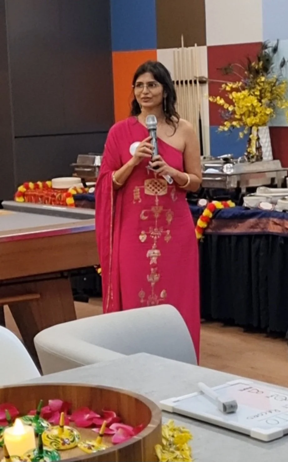 A woman in a pink saree holding a microphone, standing in front of a decorated table with flowers and candles, with a colorful mural and floral arrangements in the background.