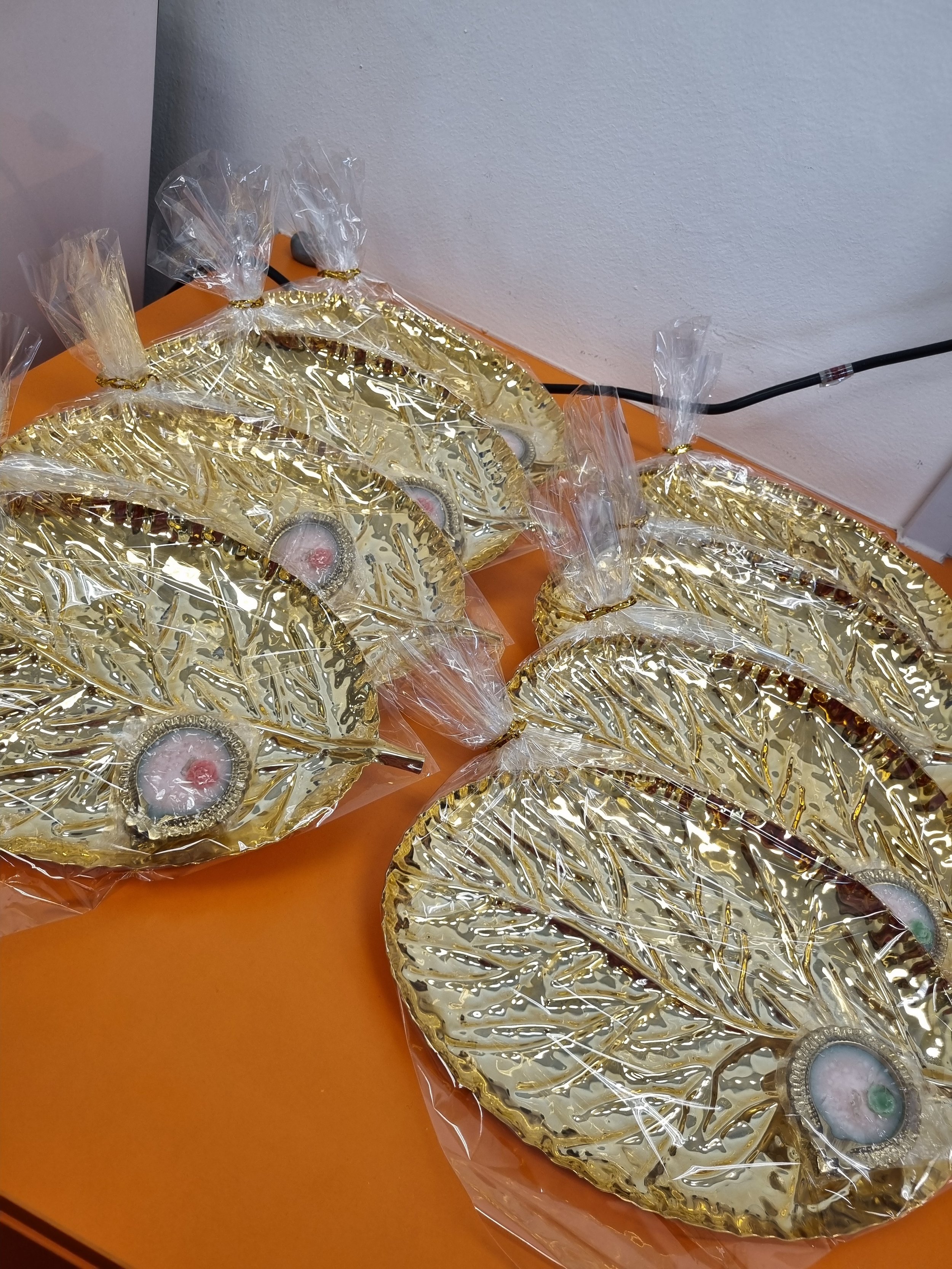 Gold-colored decorative crescent-shaped ornaments wrapped in clear plastic and tied with gold twist ties, placed on an orange surface near a white wall and black cable.