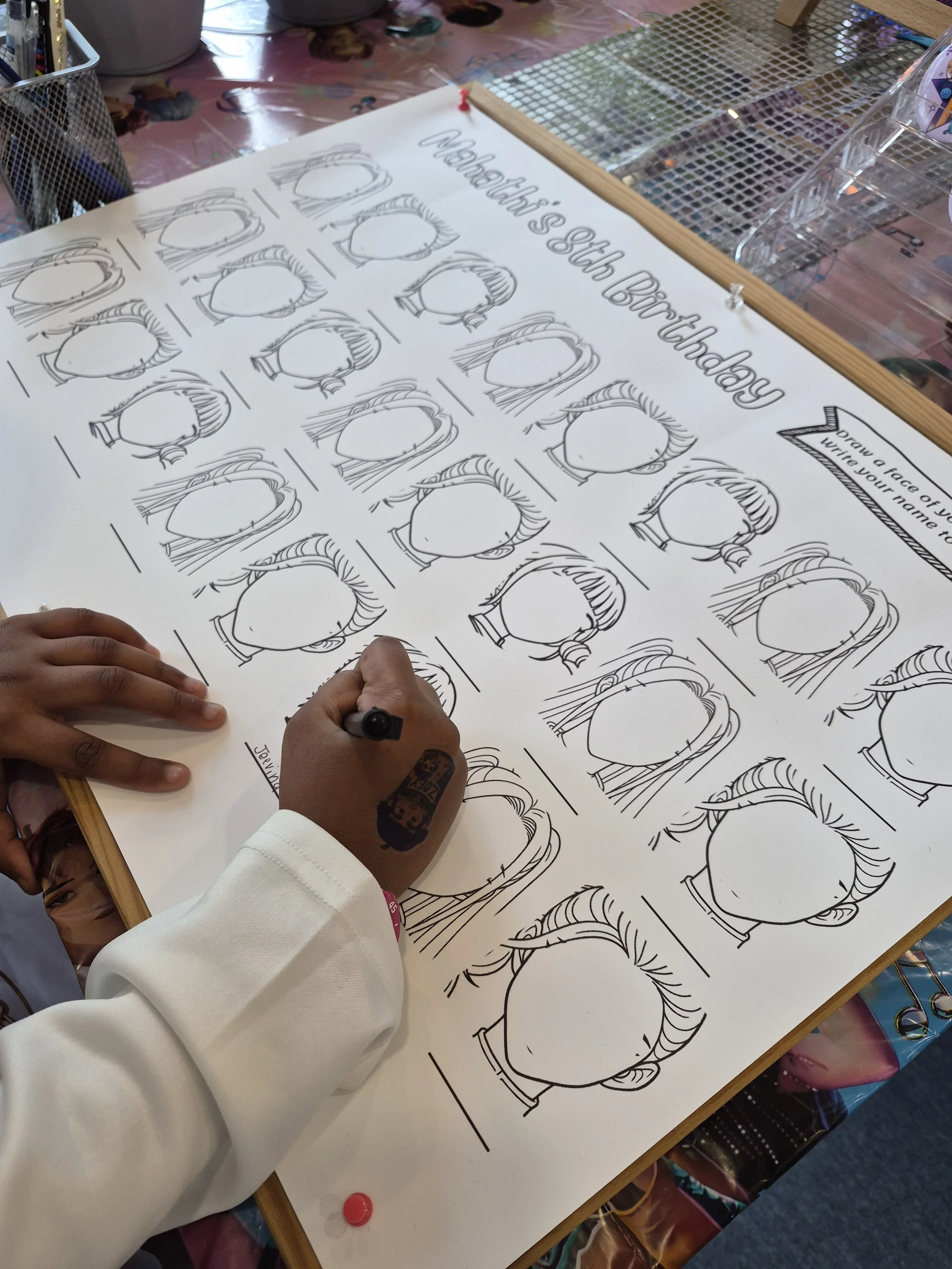 A person with dark skin is drawing a face on a large white poster with black outline sketches of different hairstyles labeled for a birthday activity, titled 'What hairstyle's kids birthday'.