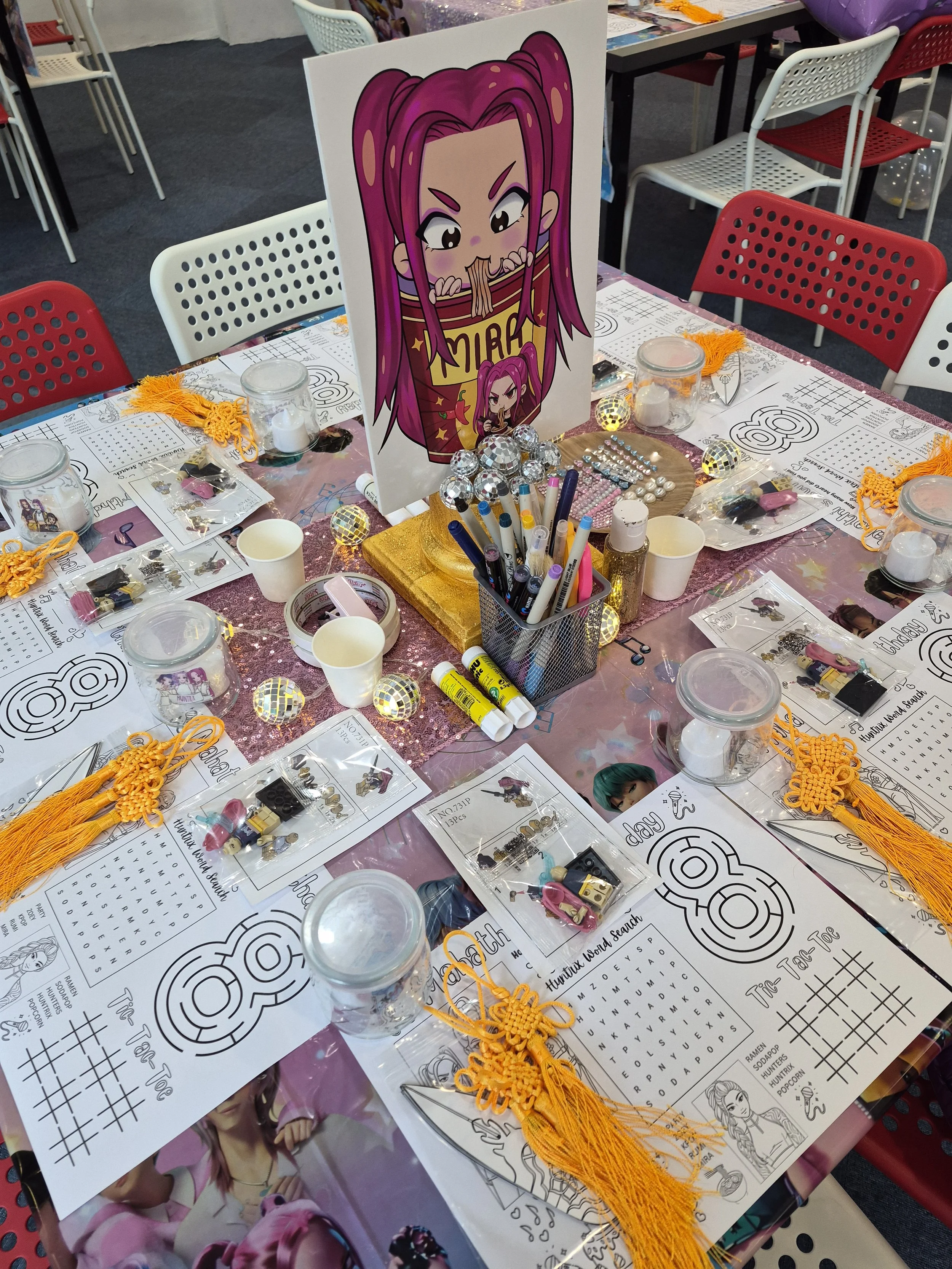 Decorative table setup for a birthday party, themed around a character with pink hair. The table has coloring pages, small jars, jewelry supplies, and orange tassel decorations. A large sign featuring the pink-haired character is at the center.