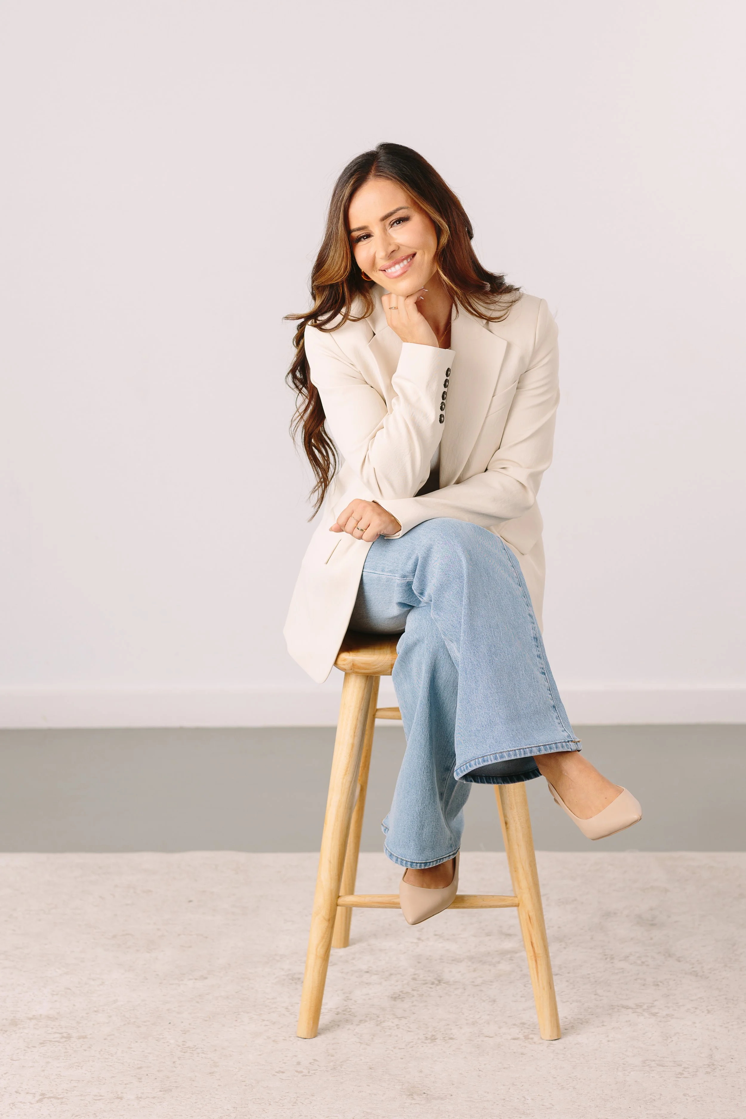 Woman with long wavy brown hair and light skin sitting on a wooden stool, smiling, wearing a cream blazer, light blue jeans, and beige heels, in front of a plain white wall.