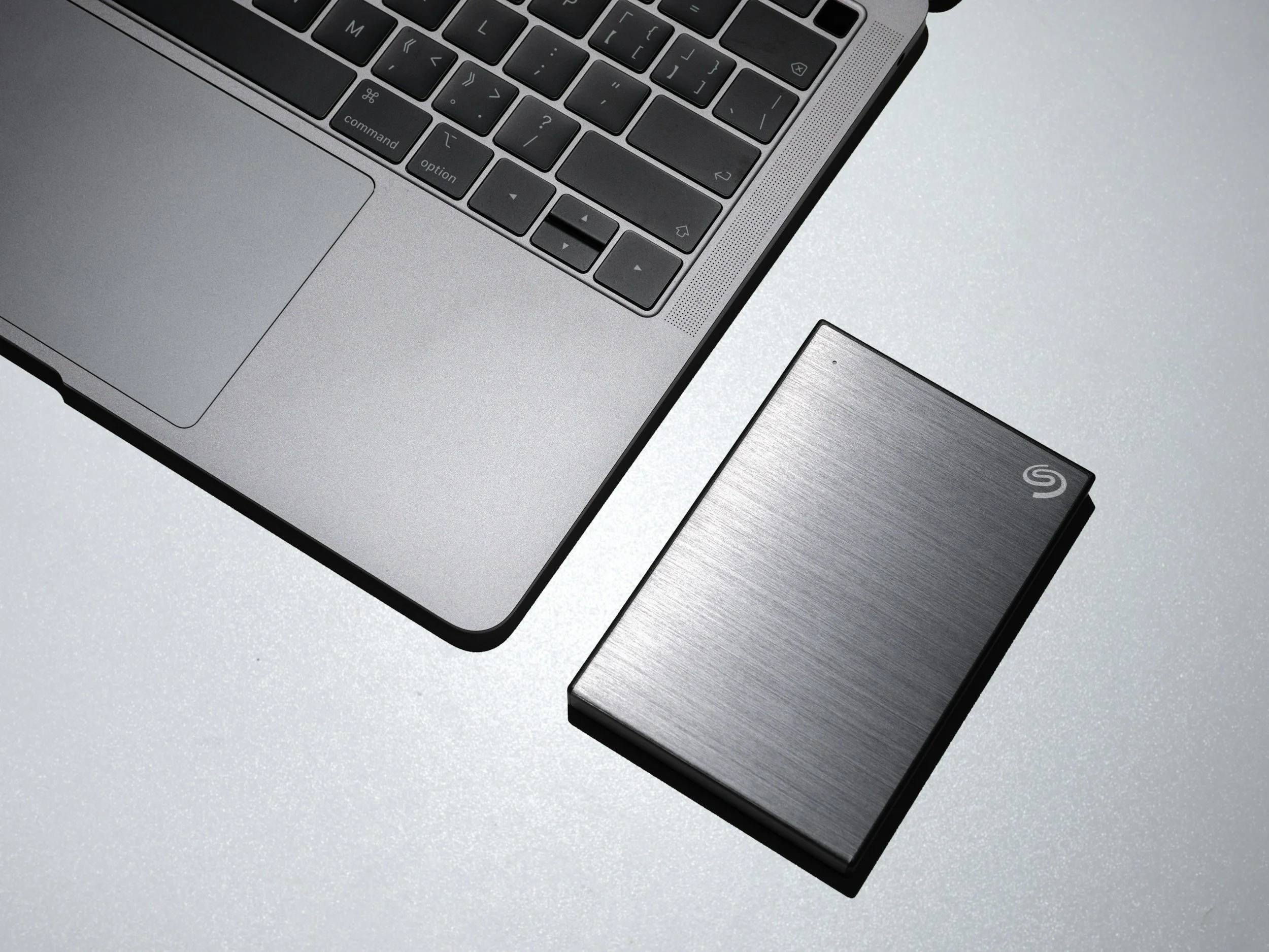 Close-up of a silver laptop keyboard and trackpad next to a black external hard drive with a brushed metal finish.