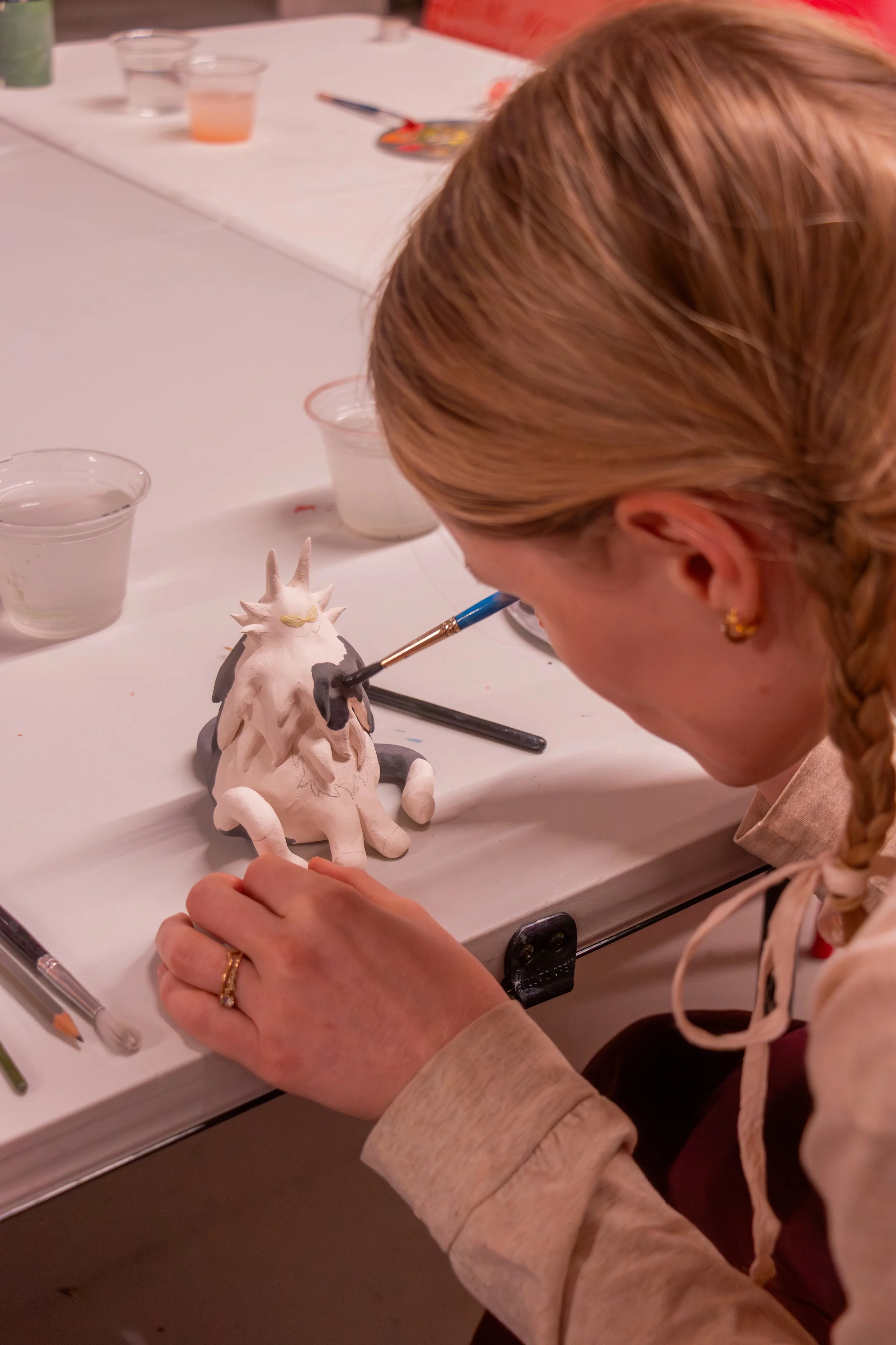 A person with reddish-blonde hair painting a small, ceramic unicorn figurine with black and yellow details on a white table in an indoor setting.