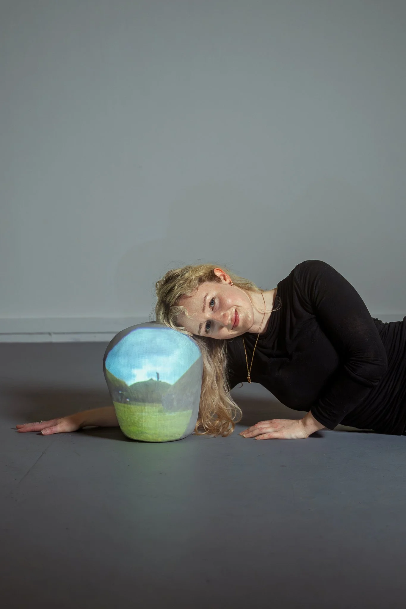 A woman with blond curly hair lying on a gray floor, resting her head on a globe-shaped paper mache object painted with landscape scenery, smiling and looking at the camera, with a light gray wall in the background.