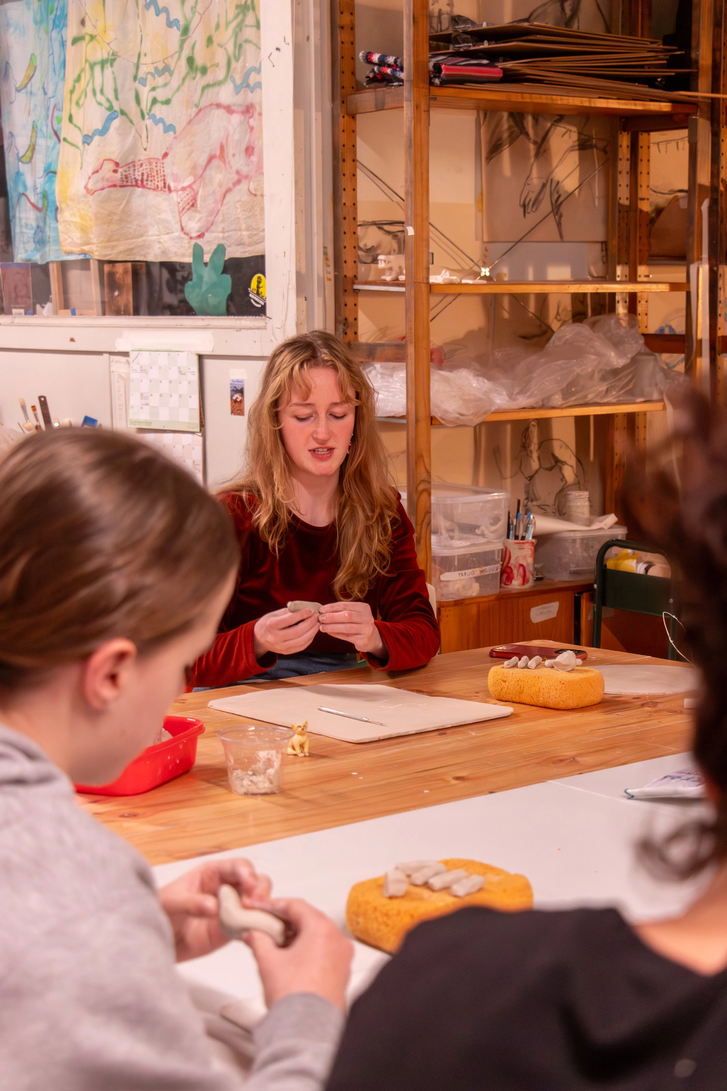 A woman is sitting at a wooden table with children, engaged in a clay modeling activity. The table has a sponge, a tray, and small clay pieces. In the background, there are shelves filled with art supplies, and a colorful, abstract painting is visible on the wall.