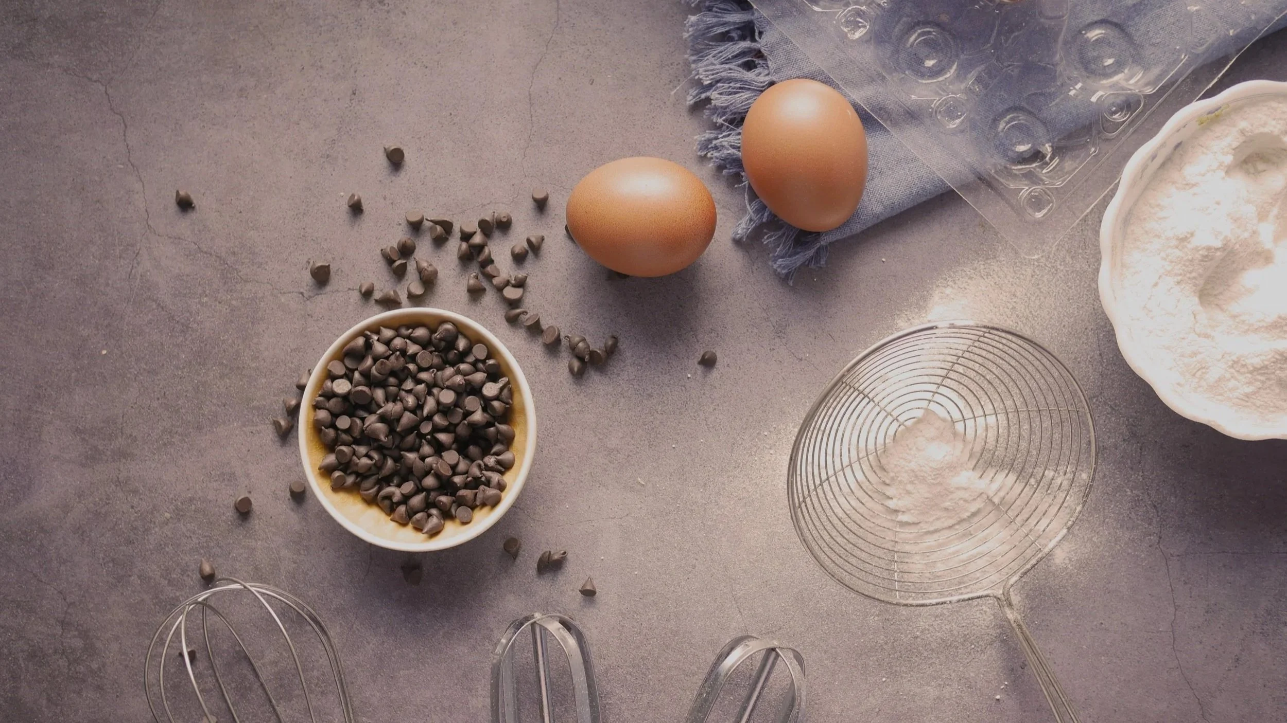 Baking ingredients and tools on a gray countertop including eggs, chocolate chips, flour, a bowl of flour, a pastry cutter, a whisk, and a spatula.