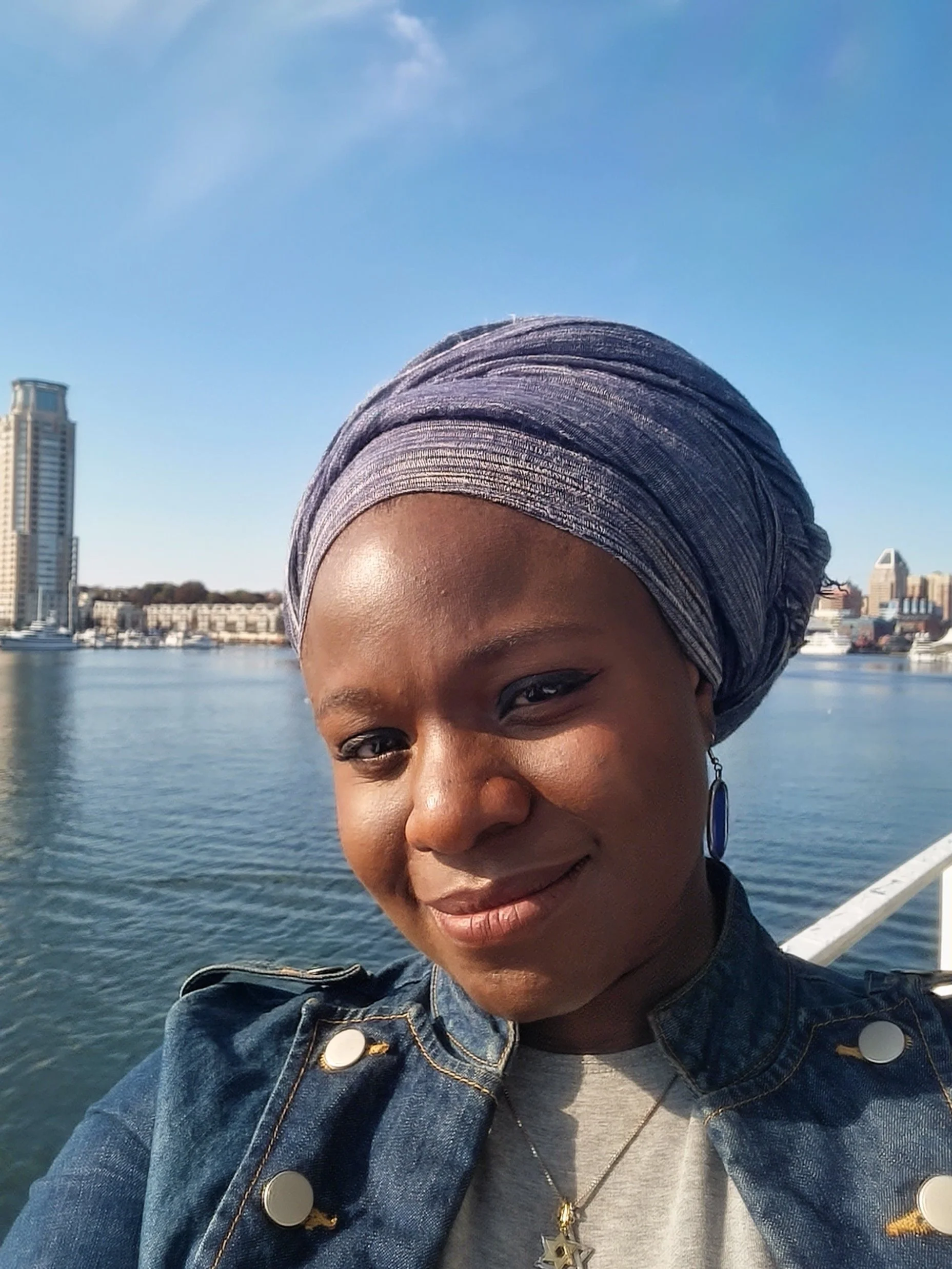 A woman smiling taking a selfie outdoors near a body of water with city buildings in the background, wearing a grey headwrap, denim jacket, and a necklace with a star pendant.