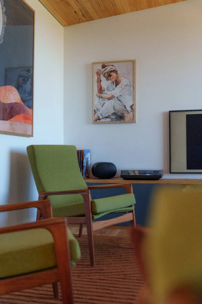 Living room with wood-paneled ceiling, green armchairs, a sofa, a coffee table, a bookshelf, a television, artwork on the walls, and a large window with a view of trees and the ocean.