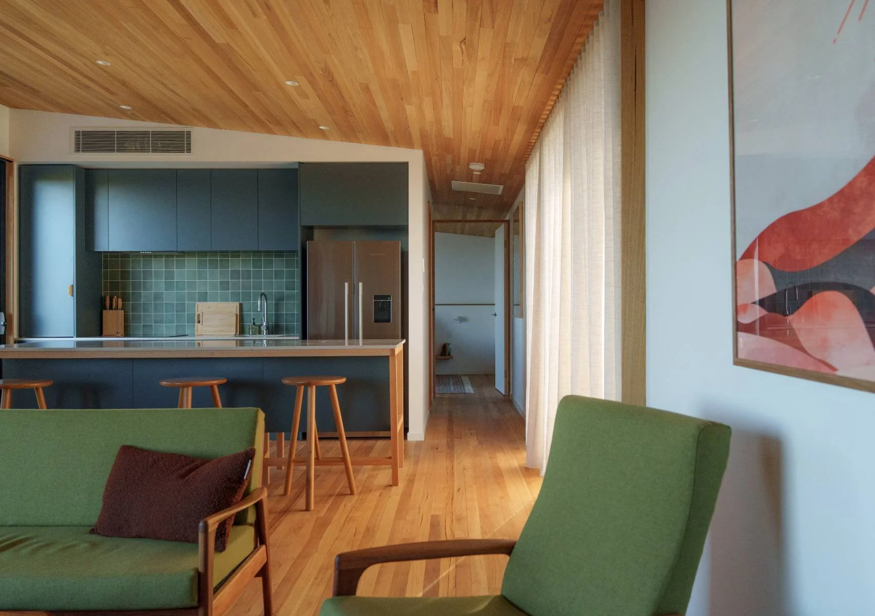 Modern kitchen with wooden ceiling and floor, large windows with a view of trees and sky, and a white kitchen island with three wooden stools.