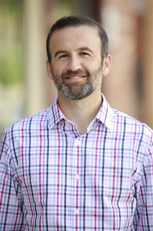 Man with short dark hair and beard smiling, wearing a multicolored checkered shirt, outdoors with blurred background.