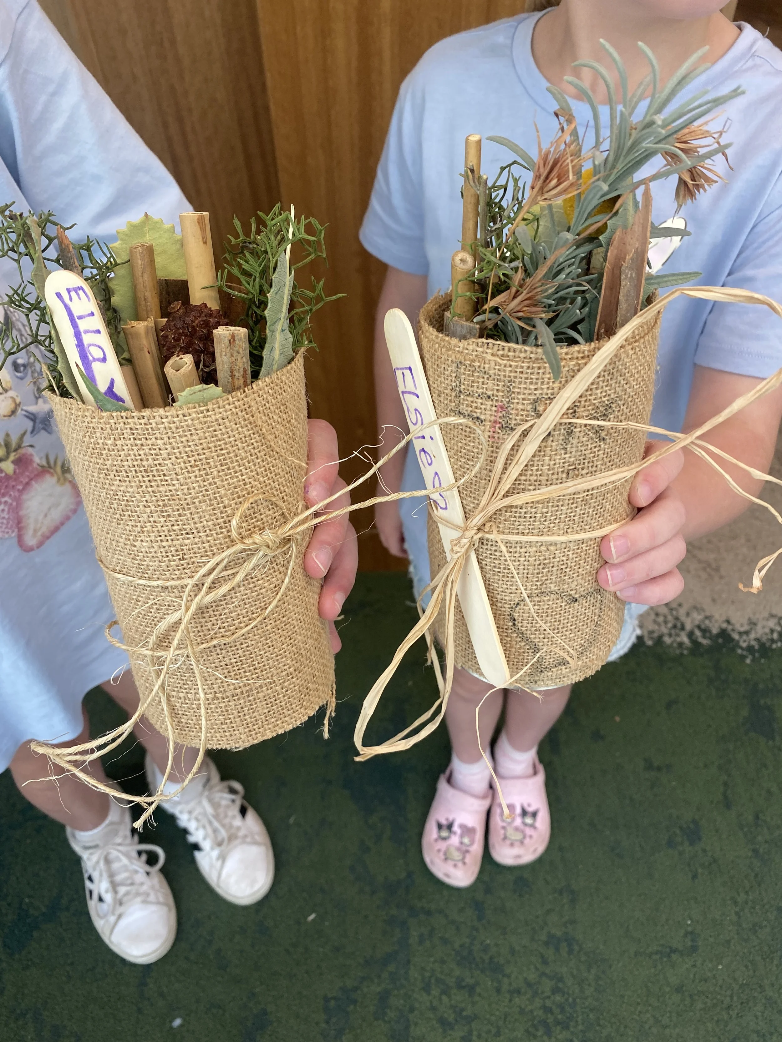 Two people hold burlap-wrapped plant arrangements decorated with dried foliage, wooden stakes, raffia, and handwritten labels.