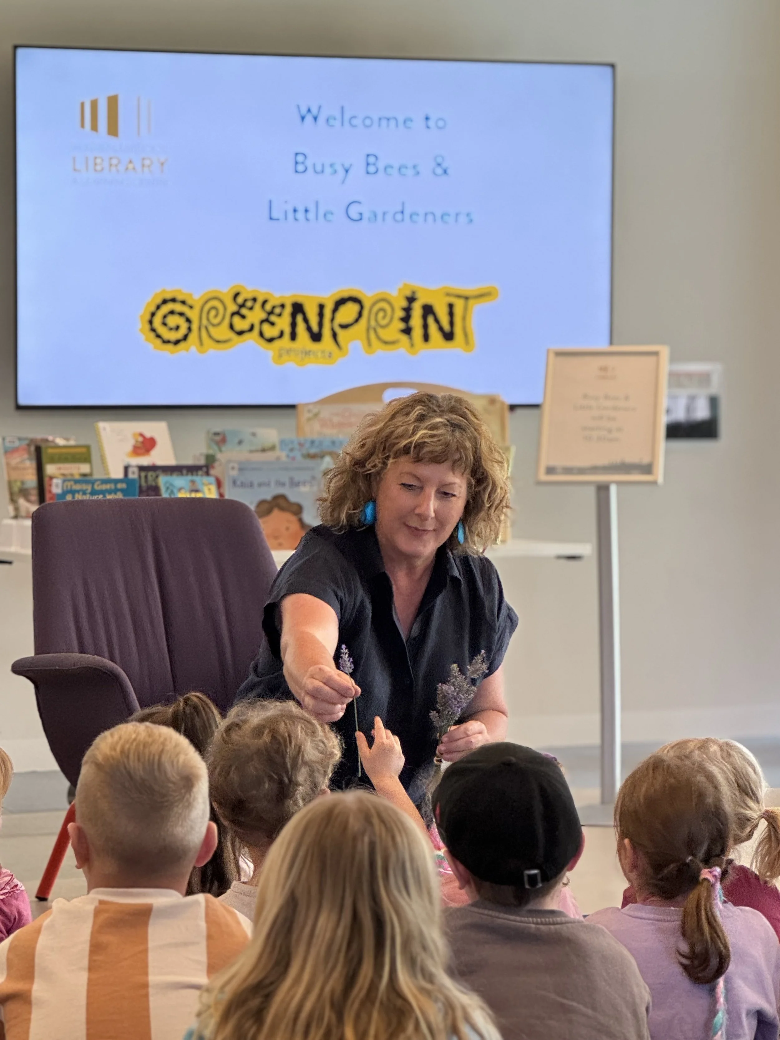 Librarian leading a children's storytime at a library beneath a screen reading “Welcome to Busy Bees & Little Gardeners.”