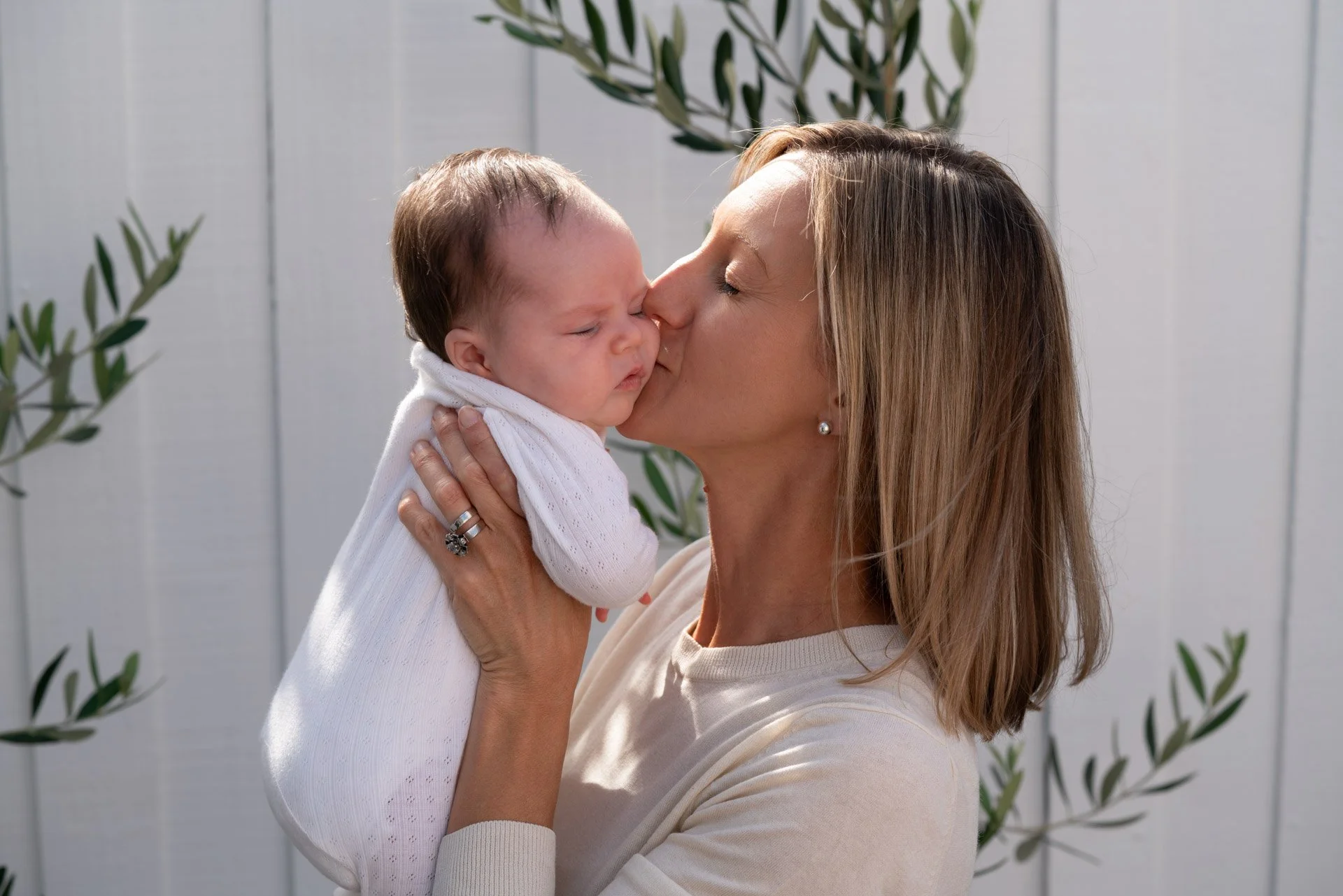 A woman with blonde hair giving a gentle kiss to a baby she is holding, with a light-colored background and green branches.