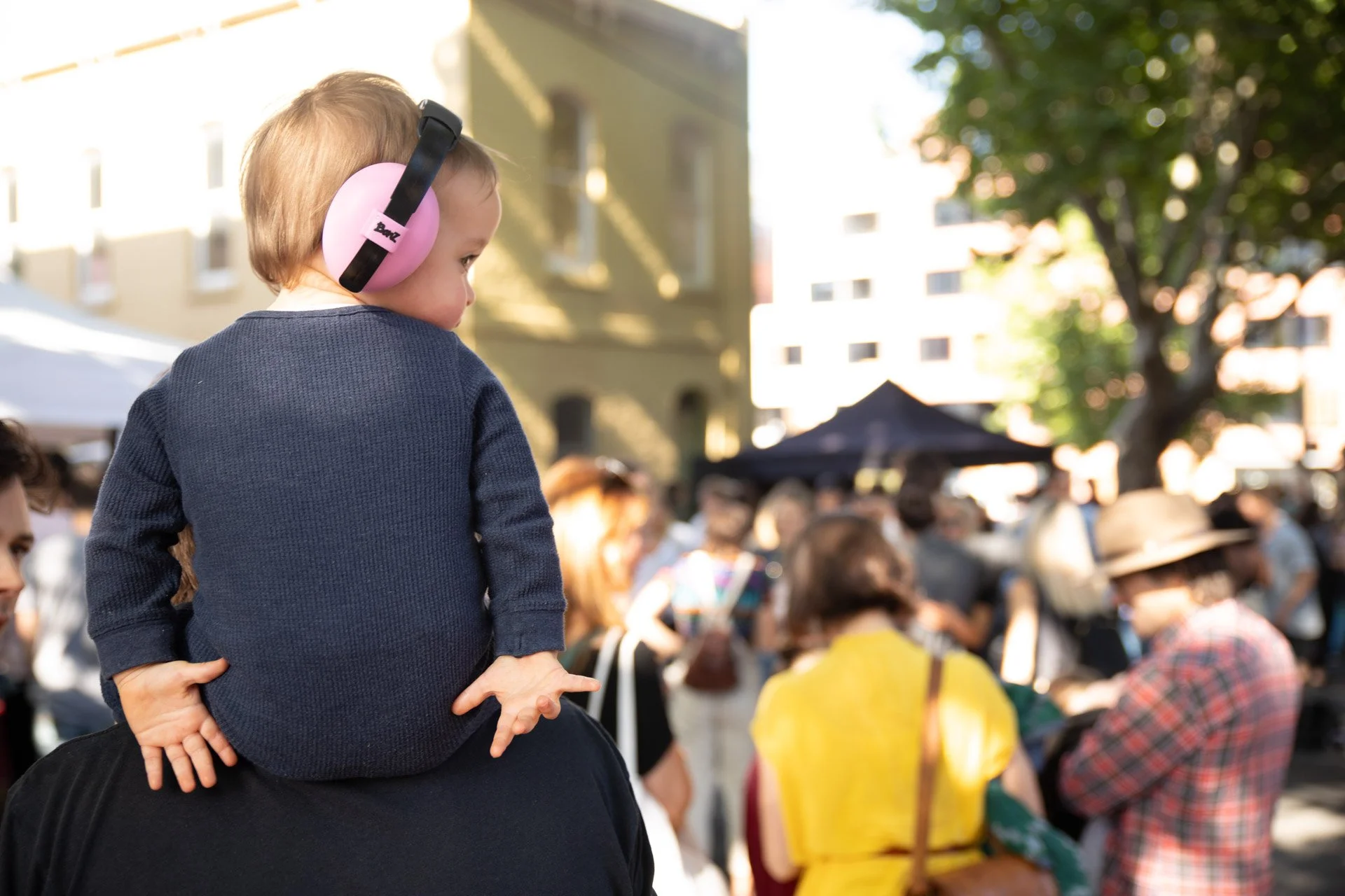 A young child with pink headphones sitting on an adult's shoulders in a crowded outdoor music festival (Peel St Festival) with people, tents, trees, and buildings in the background.