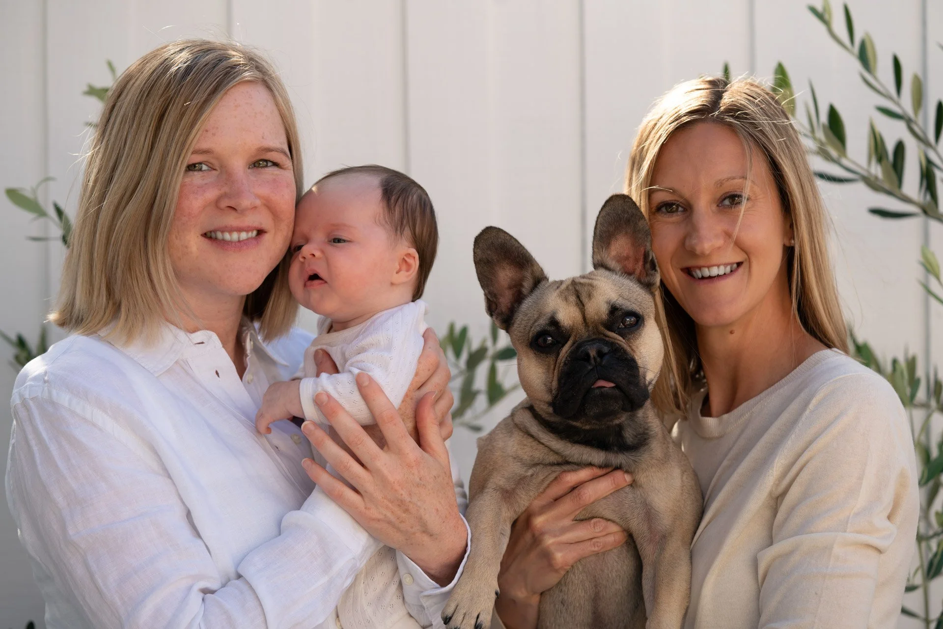 Two women smiling and holding a baby and a French Bulldog in front of a white fence with green foliage.