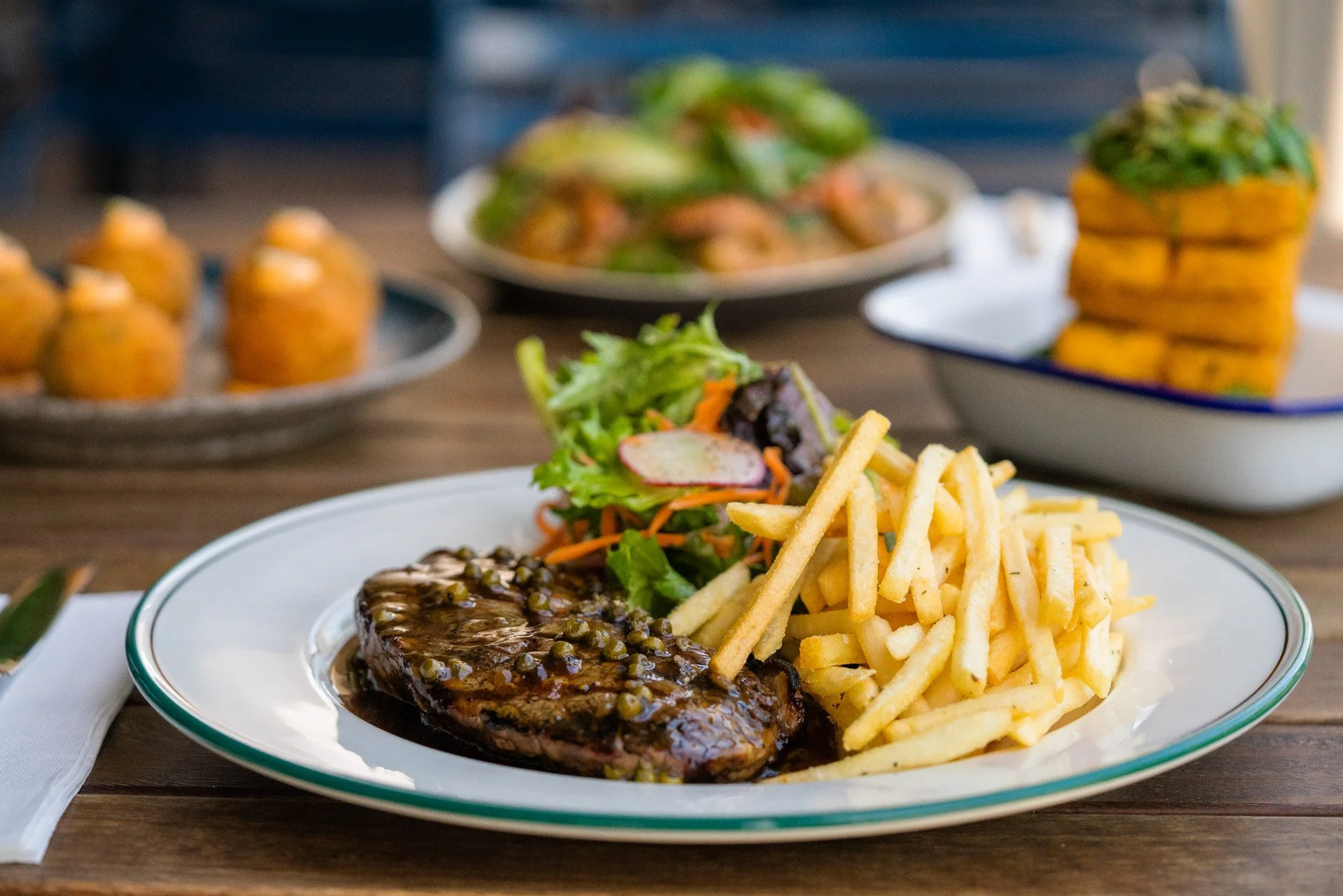 Plate with steak topped with green peppercorn sauce, French fries, and a side salad with mixed greens and vegetables. Background shows additional dishes including sliders, a salad, and stacked fried items.