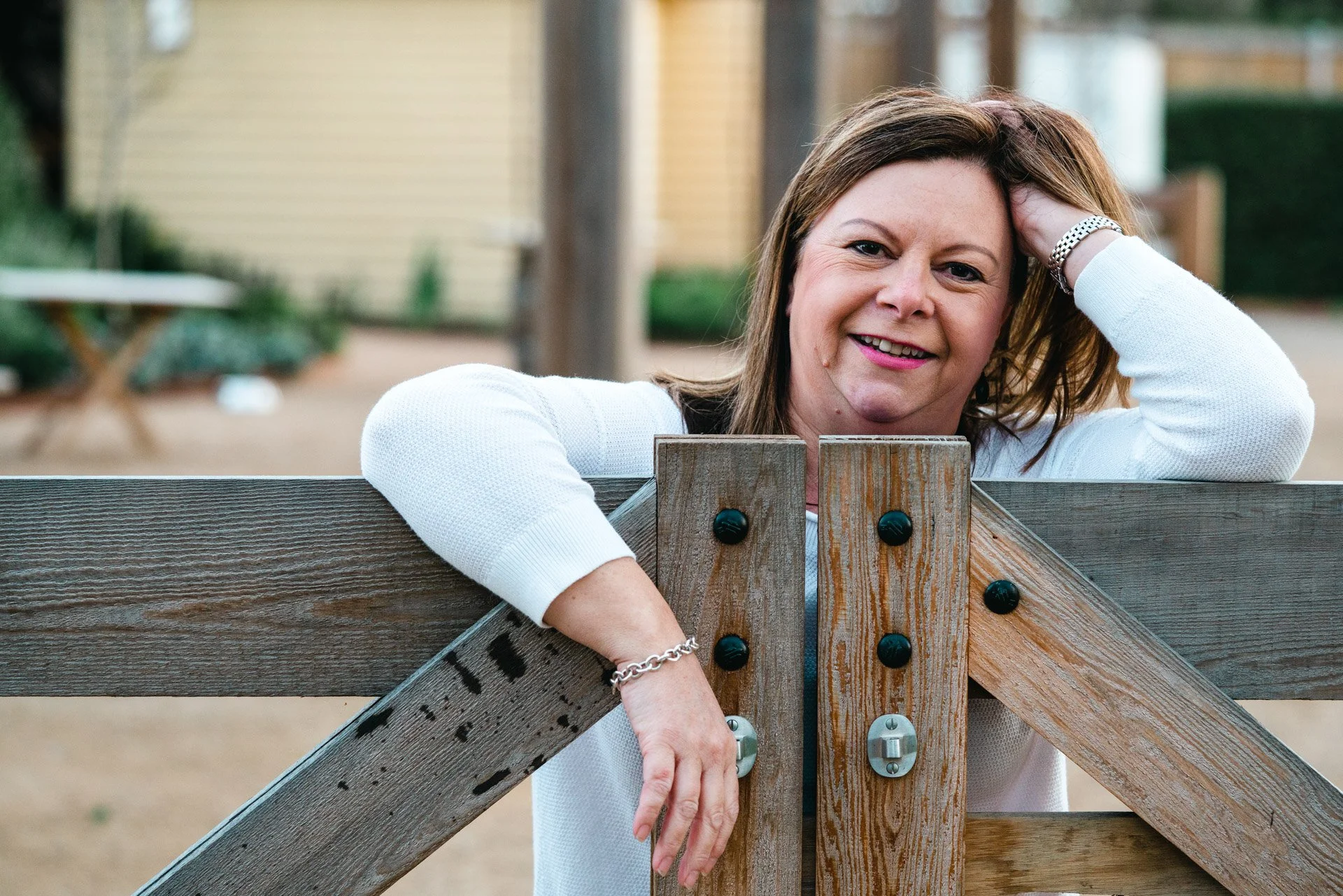 A smiling woman with shoulder-length brown hair, wearing a white sweater and silver jewelry, leaning on a wooden fence in an outdoor setting with a blurred background of trees and a building.