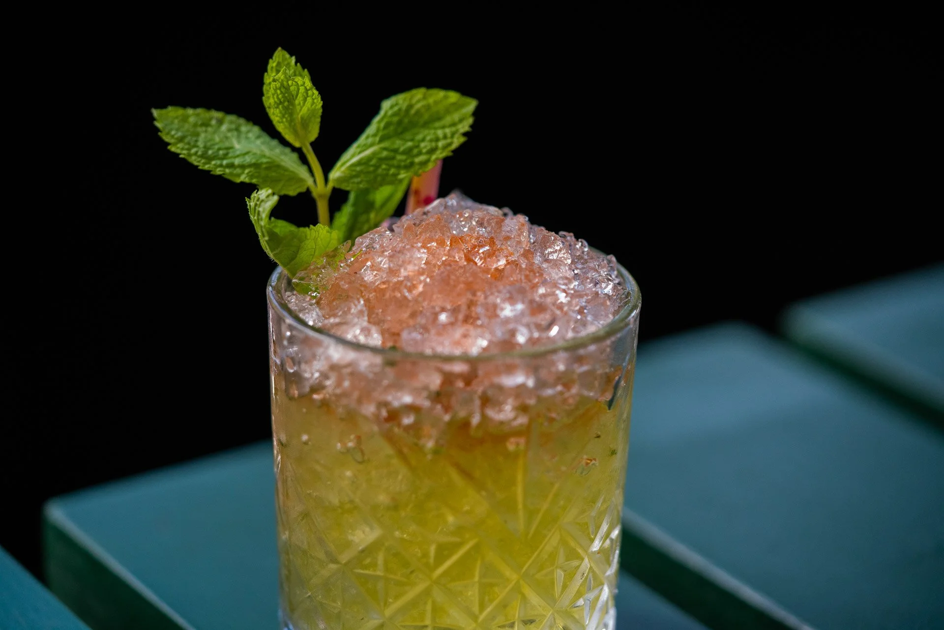 Close-up of a cocktail in a textured glass garnished with mint leaves and crushed ice against a dark background.