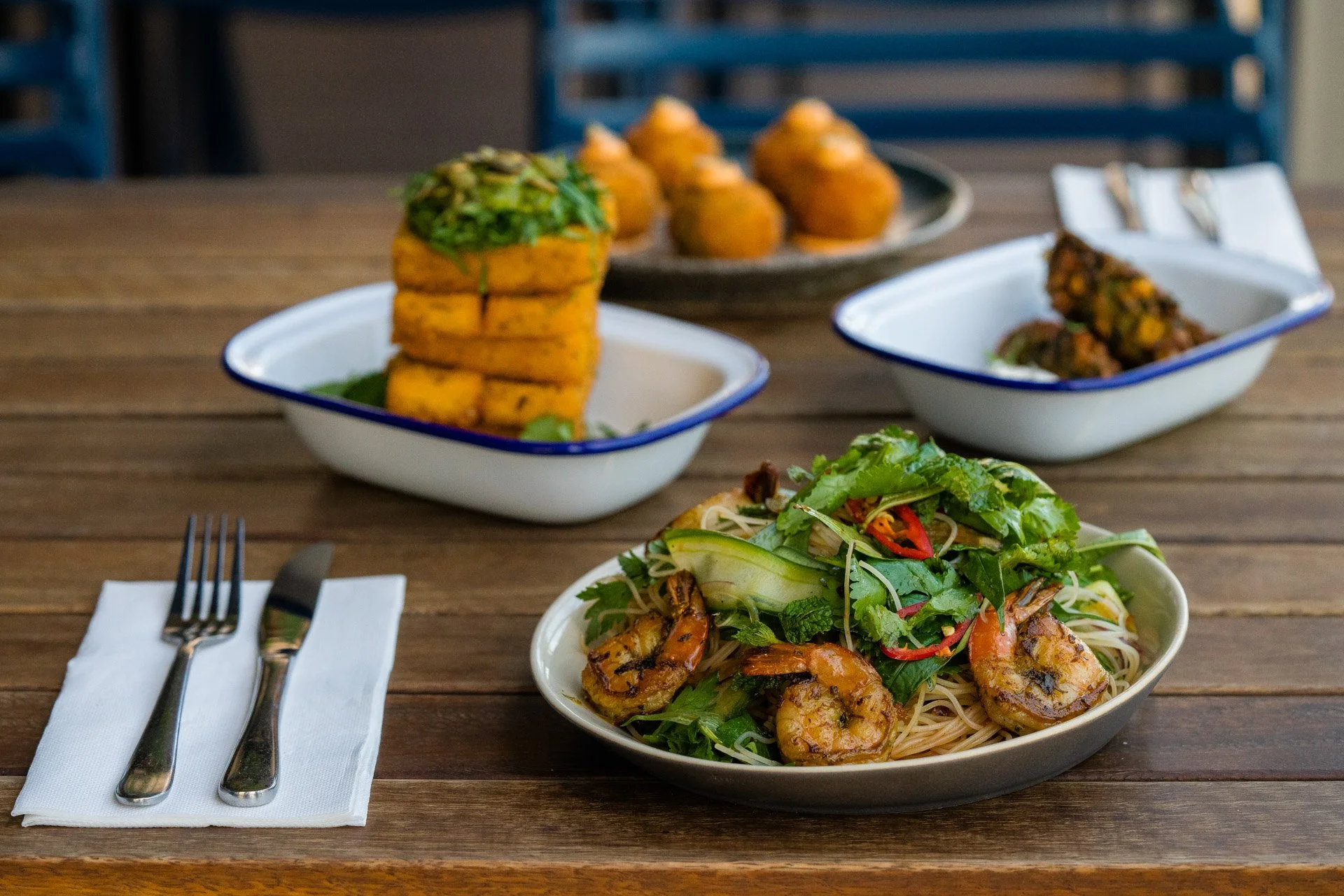 A wooden table with a dish of shrimp and noodles topped with mixed greens in the foreground, and three types of appetizer dishes in the background: fried food sticks, round filled pastries, and a piece of meat with vegetables.